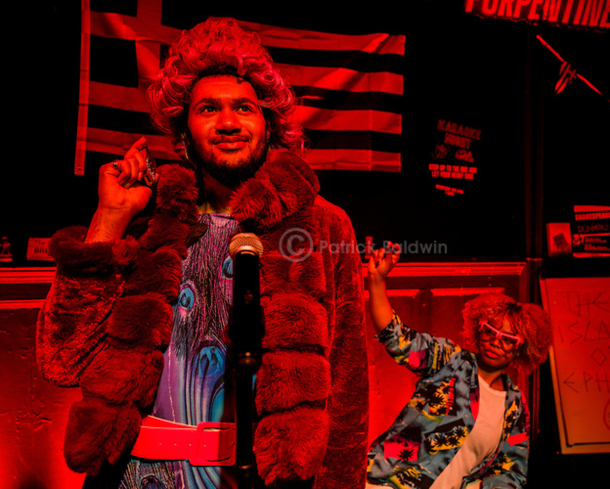 A man with a beard and curly hair dressed in a colorful outfit with a faux fur coat and wig, standing behind a microphone, with a woman with curly hair, glasses, and a tropical shirt raising her arm next to him in a room with American flags and political signs in the background, lit with red lighting.
