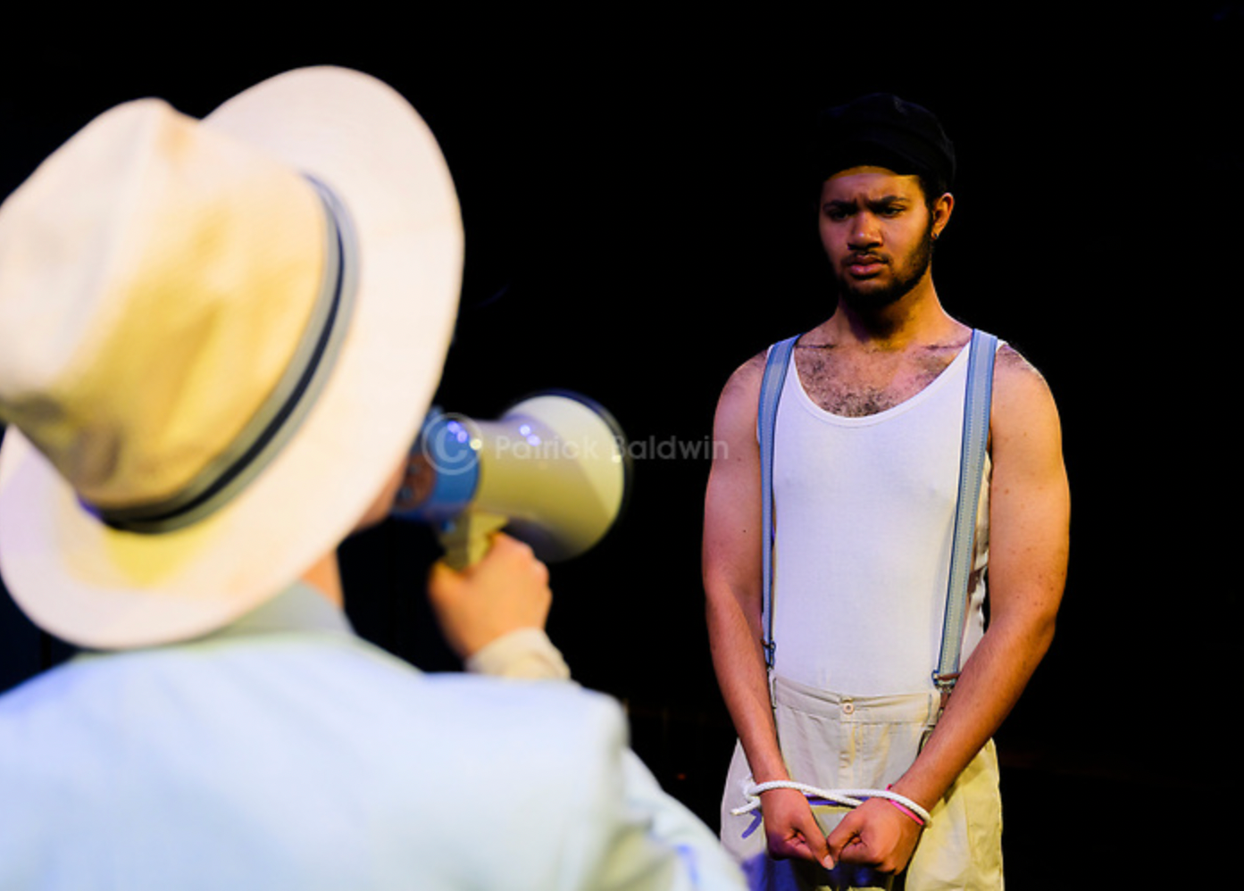 A man with a beard in suspenders and a tank top standing with hands tied, looking serious, while another person with a hat holds a megaphone towards him against a black background.