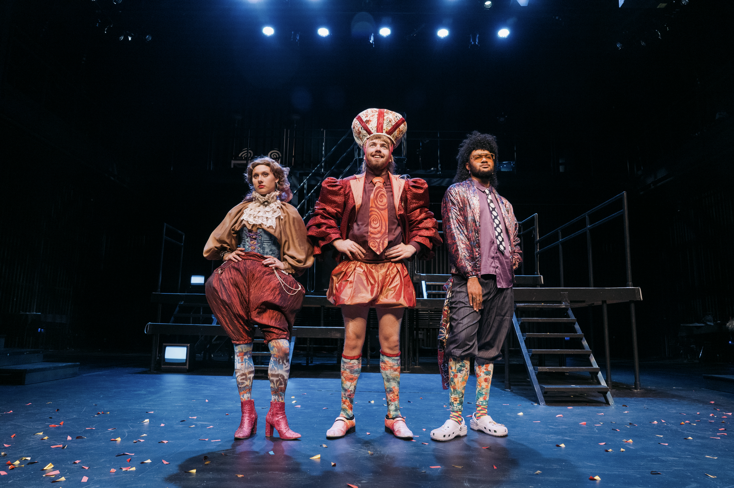 Three actors standing on stage in colorful, eccentric costumes, with a theatre set and stairs behind them, and confetti on the floor.