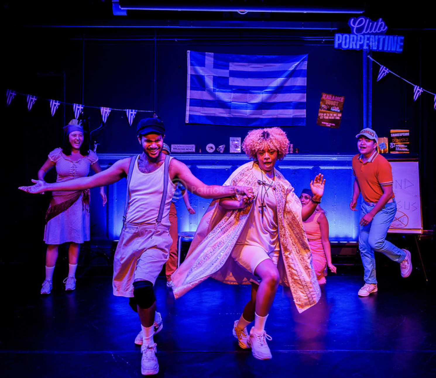 Group of young actors performing on stage with Greek flags and a sign that reads 'Club PORPE' in the background, some smiling and dancing.