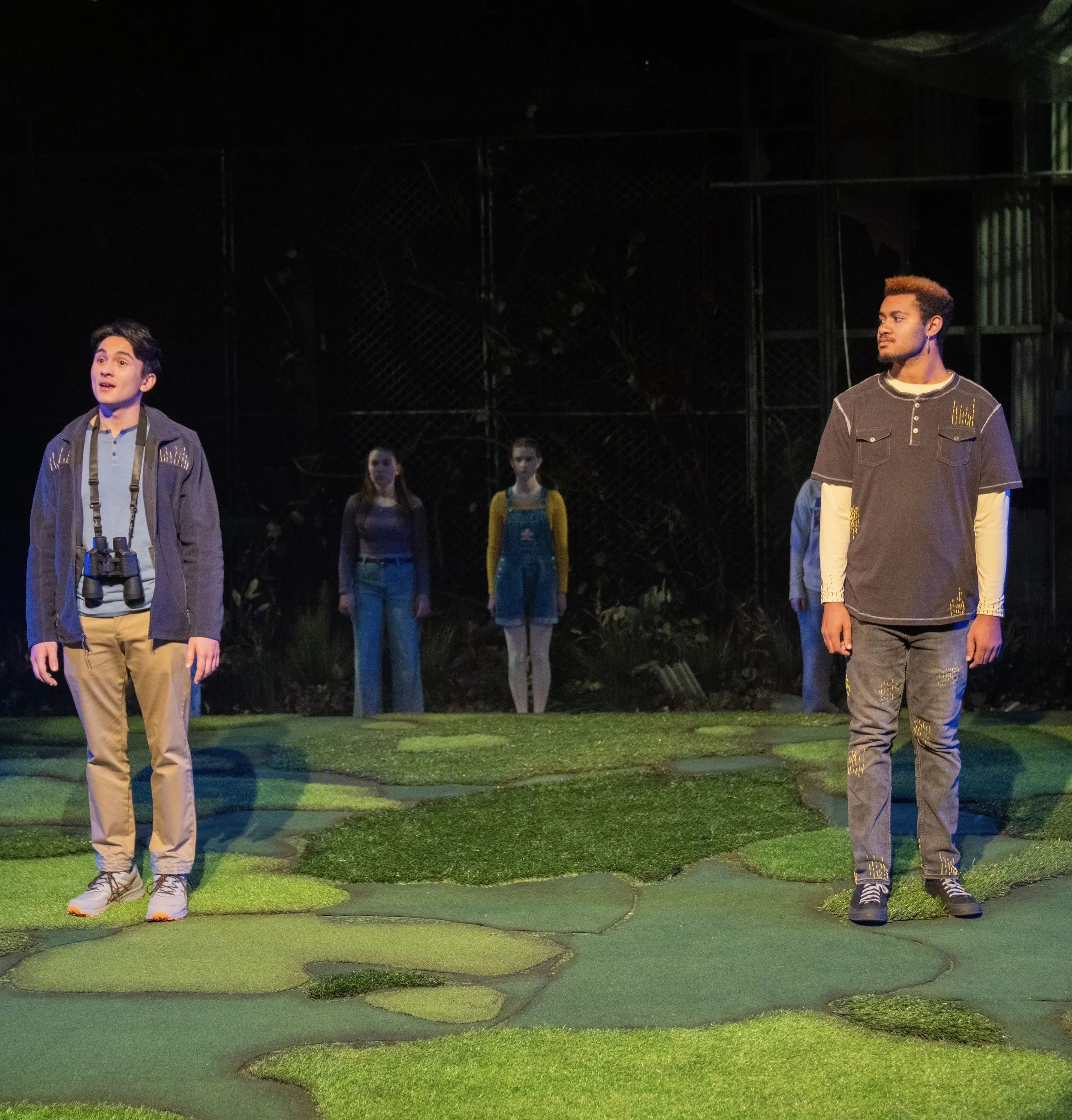 Three actors standing on a stage with a naturalistic set, two men in the foreground and two women in the background.