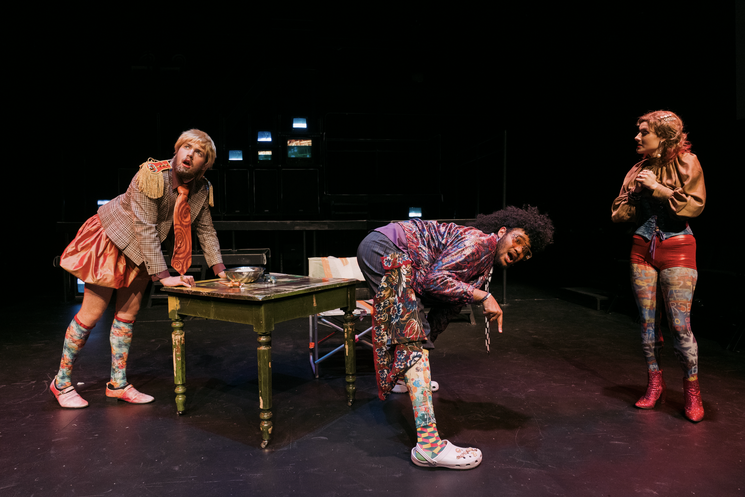 Three actors performing on stage in colorful costumes. One is leaning on a table with a surprised expression, another is crouching down pointing, and the third is standing with hands clasped, appearing engaged in a theatrical scene.