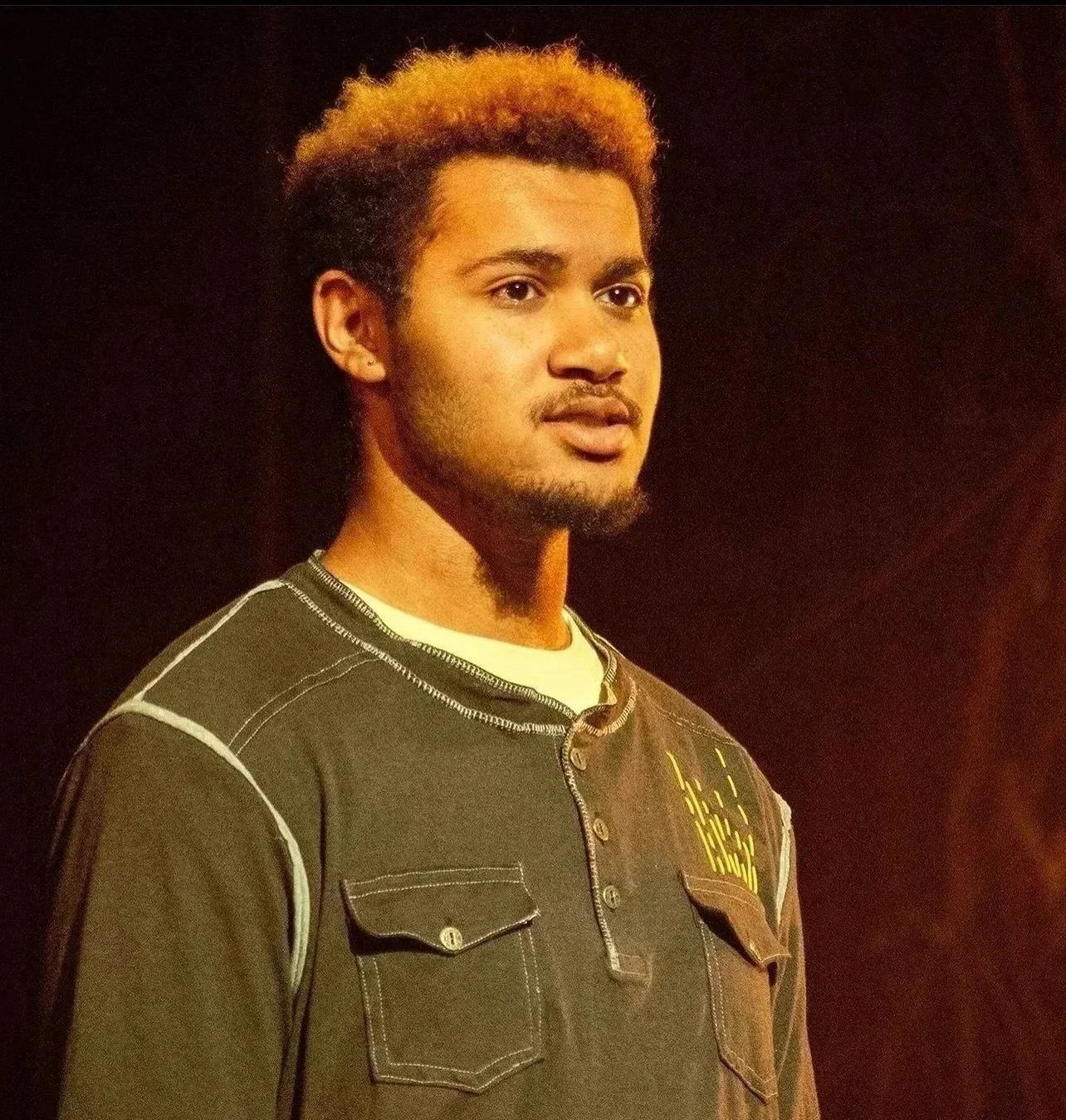 A young man with light brown skin, short curly hair with blond tips, and a goatee, wearing a brown jacket with white stitching and male shirt, standing against a dark background.