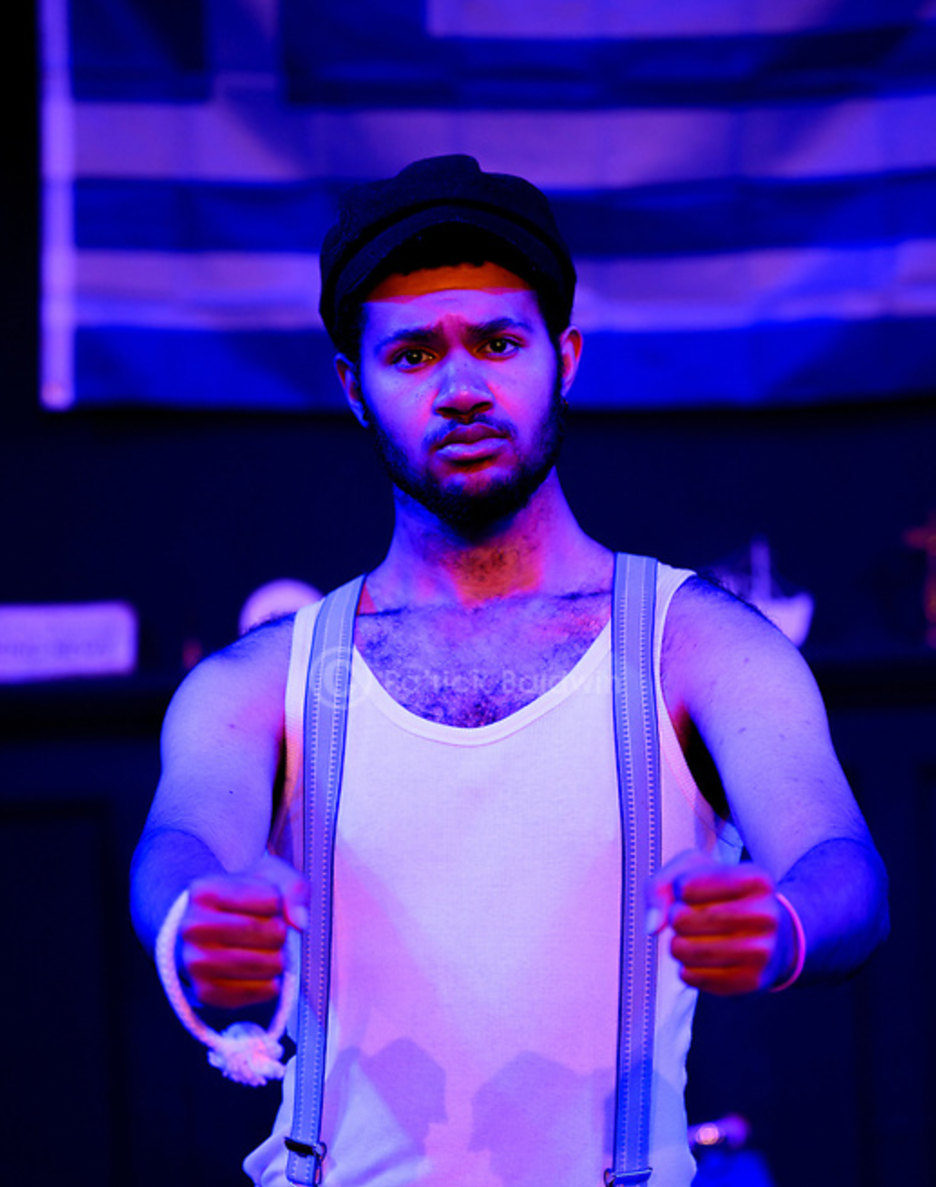 Young man with a serious expression, wearing a flat cap, tank top, and suspenders, standing in a neon-lit environment with American flag in the background.