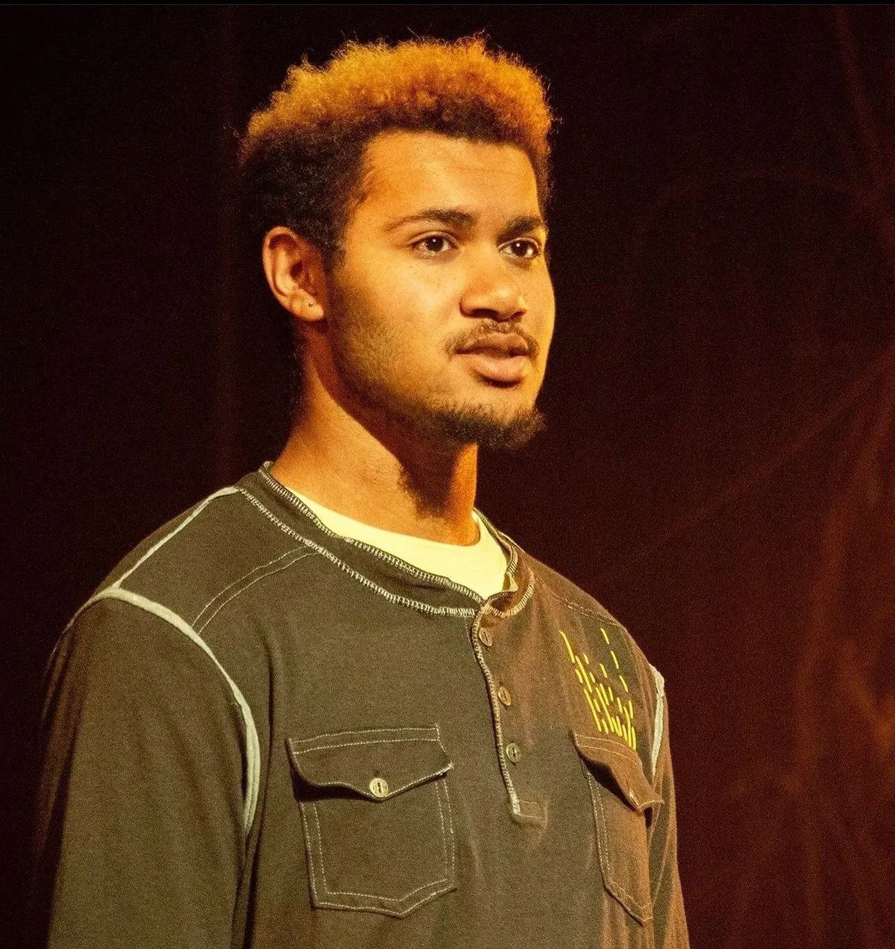Young man with curly, blonde-tipped hair, wearing a dark shirt with white stitching and chest pockets, standing against a dark background.