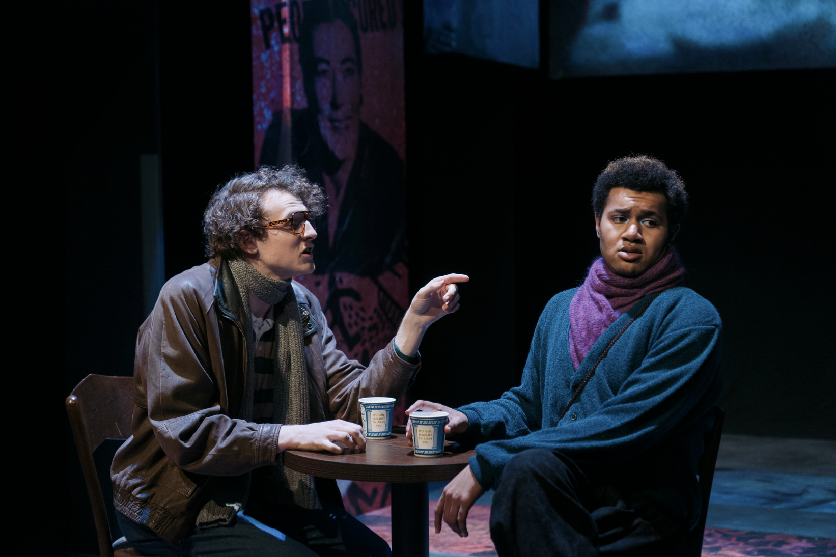 Two young men sitting at a small round table during a theatrical performance. The man on the left is speaking or arguing, wearing sunglasses, a striped shirt, and a brown jacket with a scarf. The man on the right appears to be listening with a skeptical or confused expression, dressed in a blue sweater with a purple scarf. There are two cups on the table and a large poster of a smiling woman in the background.