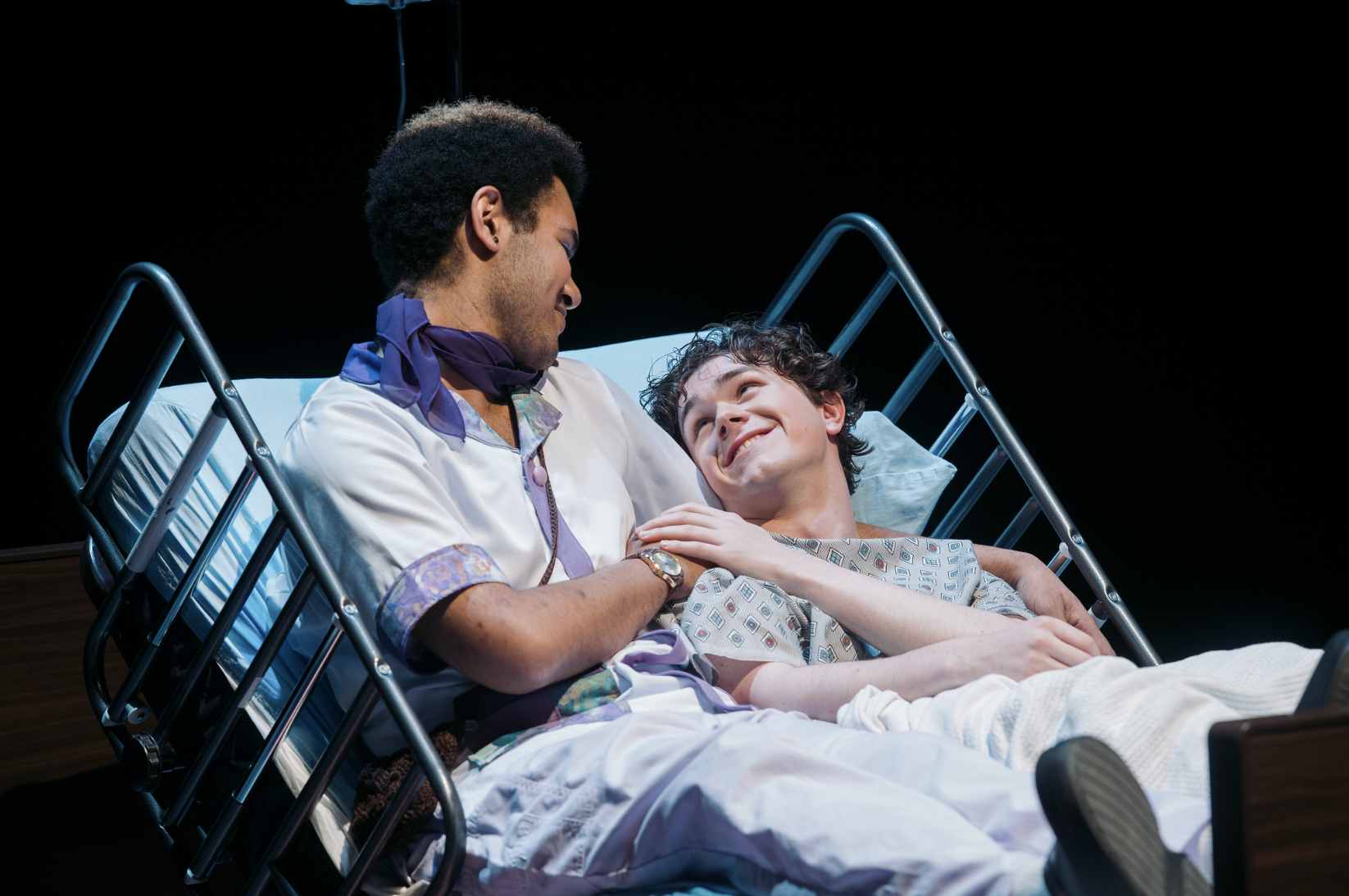 Young man lying in hospital bed, smiling and looking at a healthcare worker sitting beside him, with dark background.