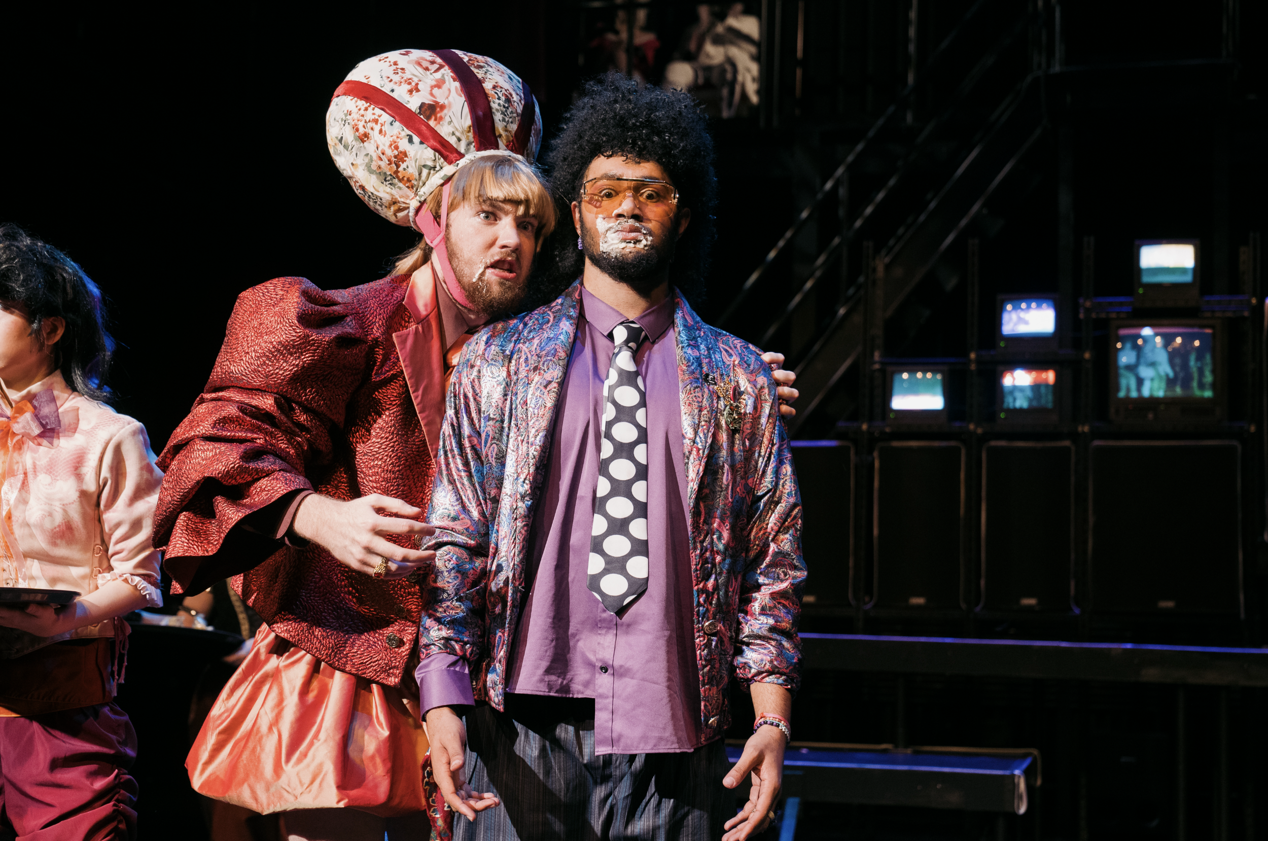 Two men performing on stage in colorful, eccentric costumes with TV screens in the background.