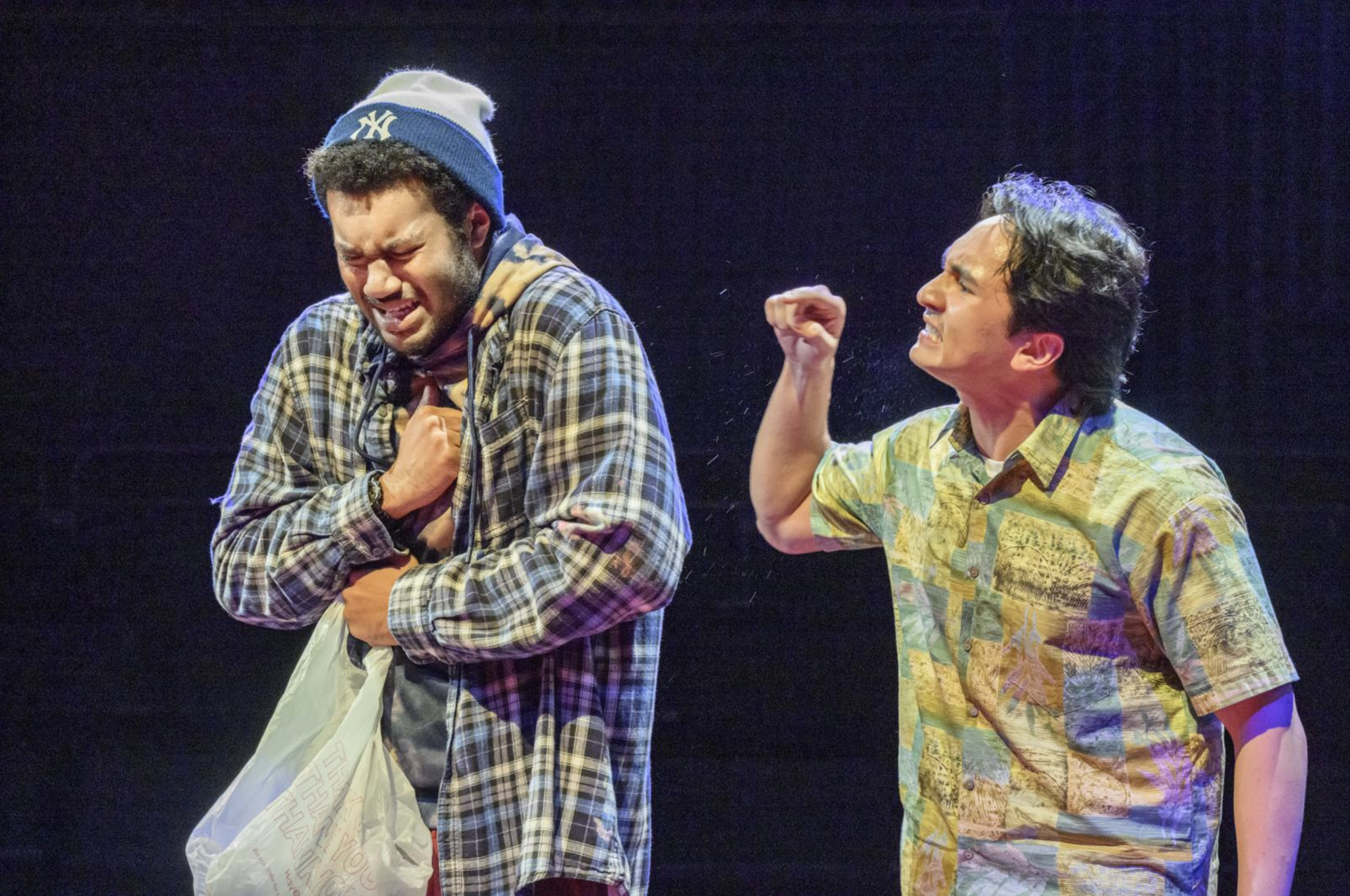 Two men are on stage, emotionally expressive; the man on the left with a beanie and plaid shirt appears to be upset, clutching a plastic bag, while the man on the right with a colorful button-up shirt is angry, raising his fist.
