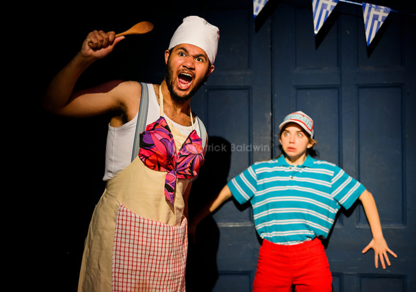 A man wearing a chef's hat, apron, and suspenders, making an angry face and holding a wooden spoon, stands next to a woman with a shocked expression, wearing a striped shirt, red shorts, and a cap, in front of a blue door with bunting flags.
