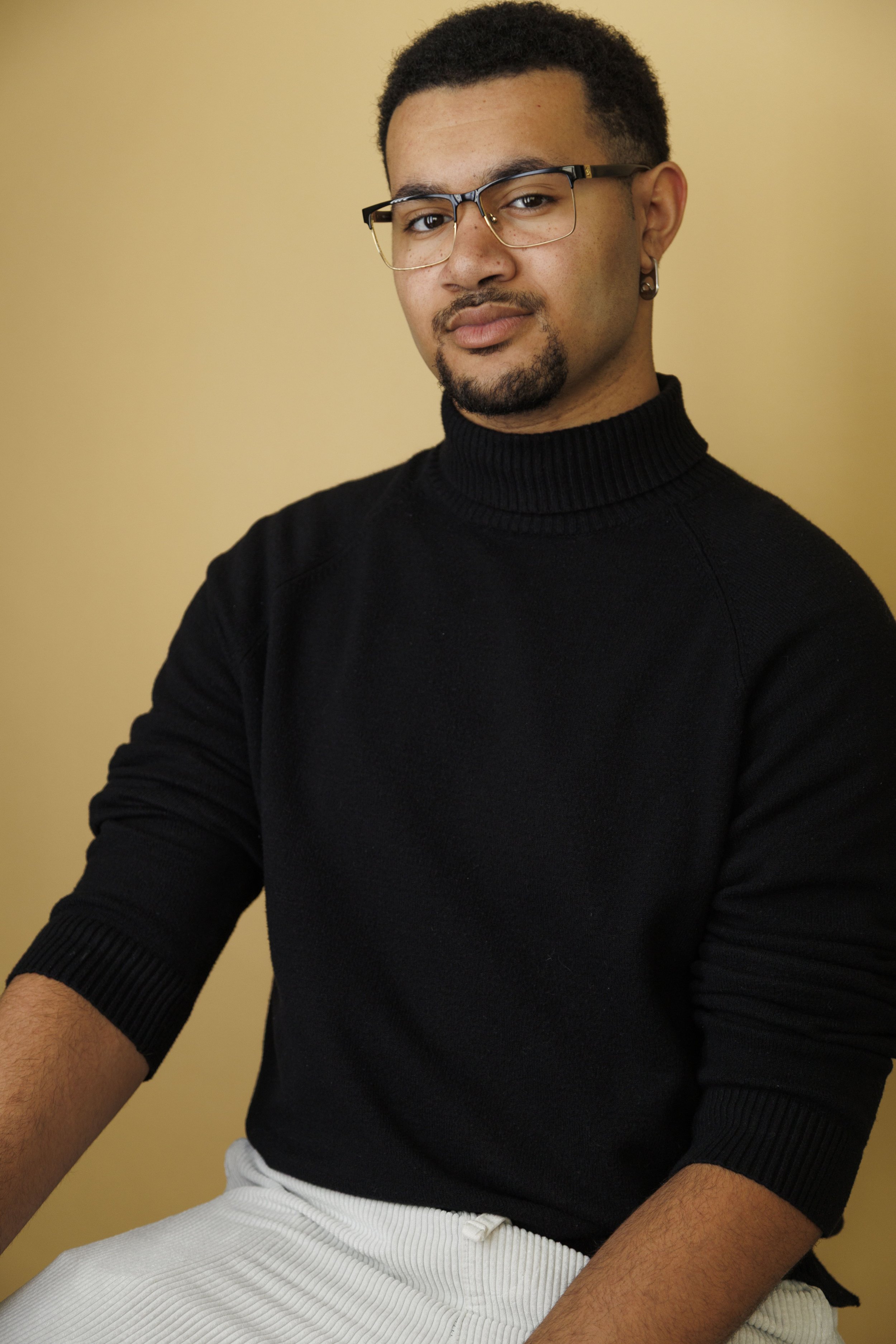 Portrait of a young man with glasses and facial hair, wearing a black turtleneck sweater, sitting against a beige background.