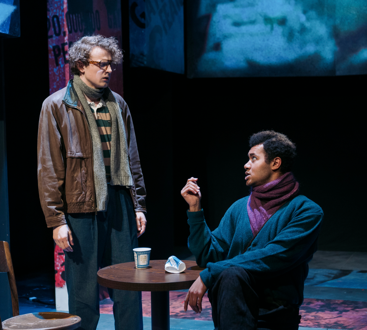 Two men having a conversation on a stage, with one sitting and the other standing. The standing man has curly hair, wears sunglasses, a leather jacket, and a scarf. The seated man has short curly hair, wears a blue coat, and a purple scarf. There is a small round table with a cup and some papers between them.