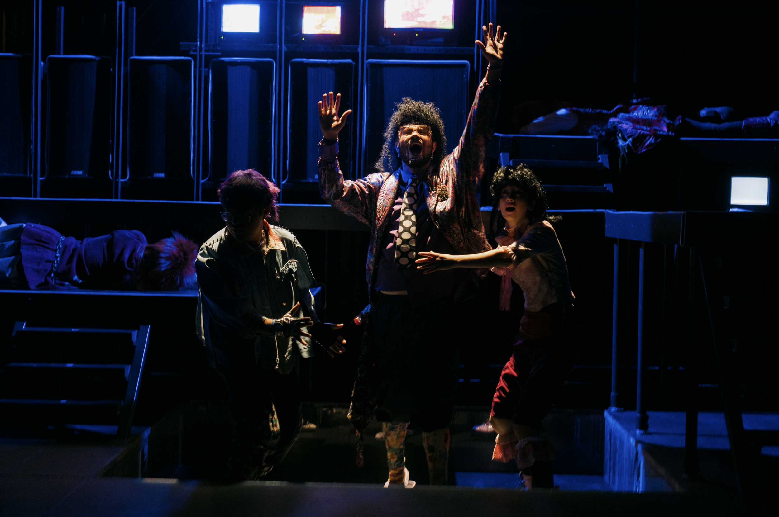 Three performers on a dark stage, with two sitting or lying on a raised platform in the background. The central performer is standing with arms raised, wearing a colorful jacket and patterned pants, singing or speaking passionately. The other two performers are interacting with him, one on his left and one on his right, dressed in vintage or eclectic costumes, with intense expressions.