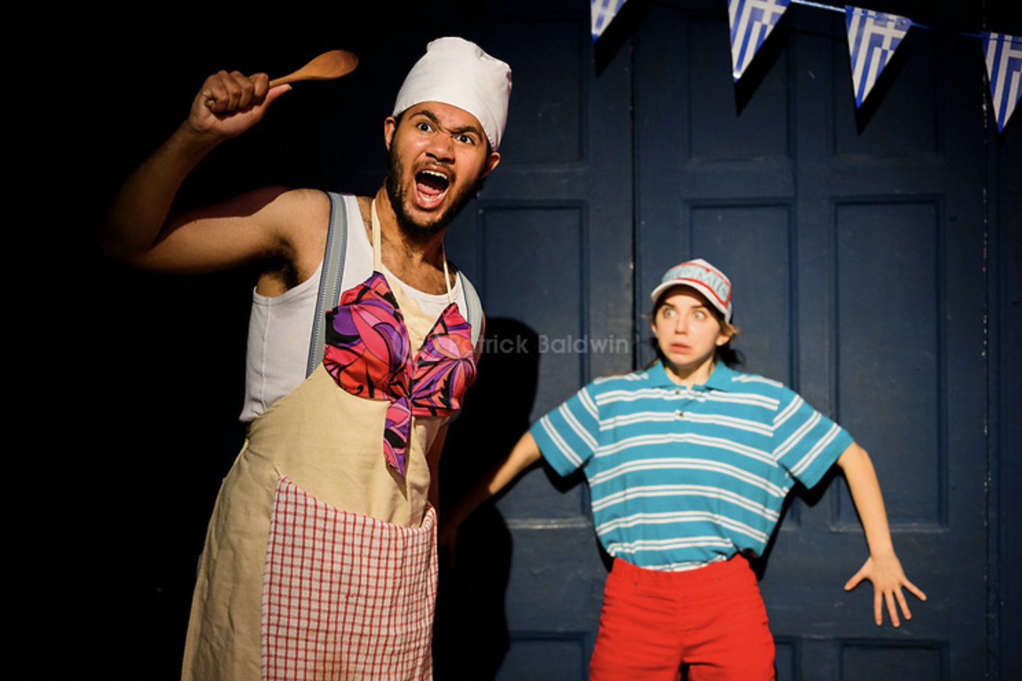 Two actors on stage, one man wearing a chef's hat and apron, holding a wooden spoon, with an angry expression; the other woman wearing a striped shirt and red shorts, with a surprised and scared expression.