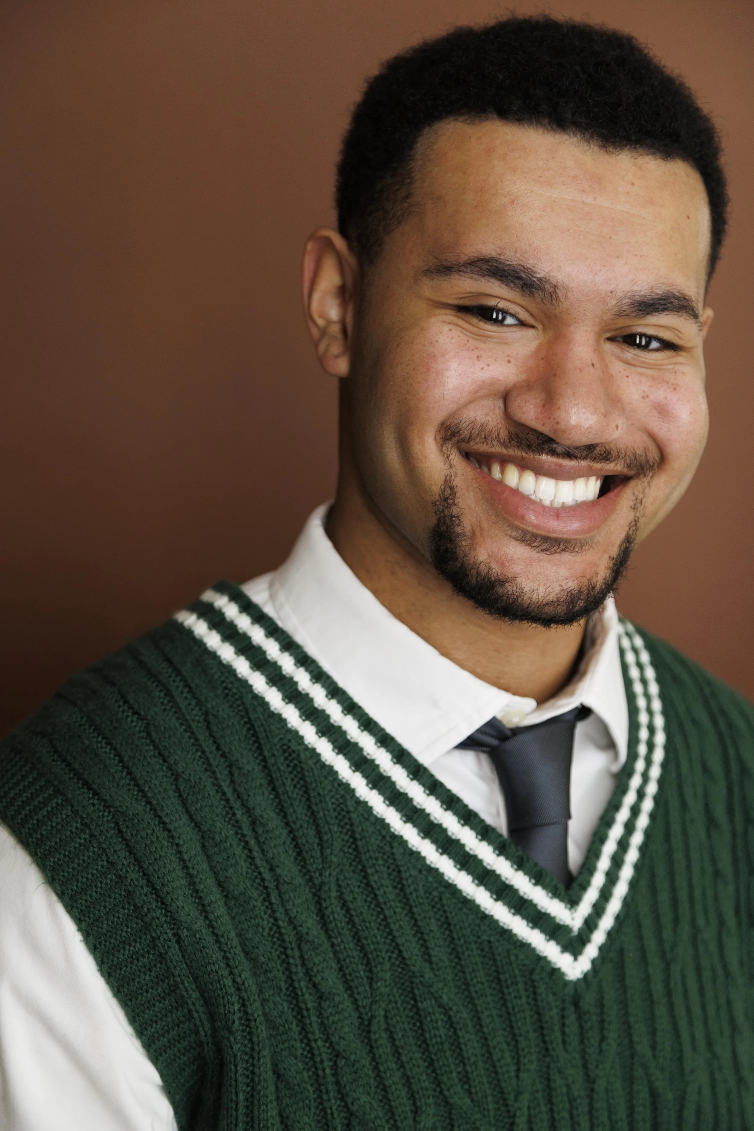 A young man with a short afro and facial hair, smiling, wearing a white shirt, a dark gray tie, and a green knitted vest, against a brown background.