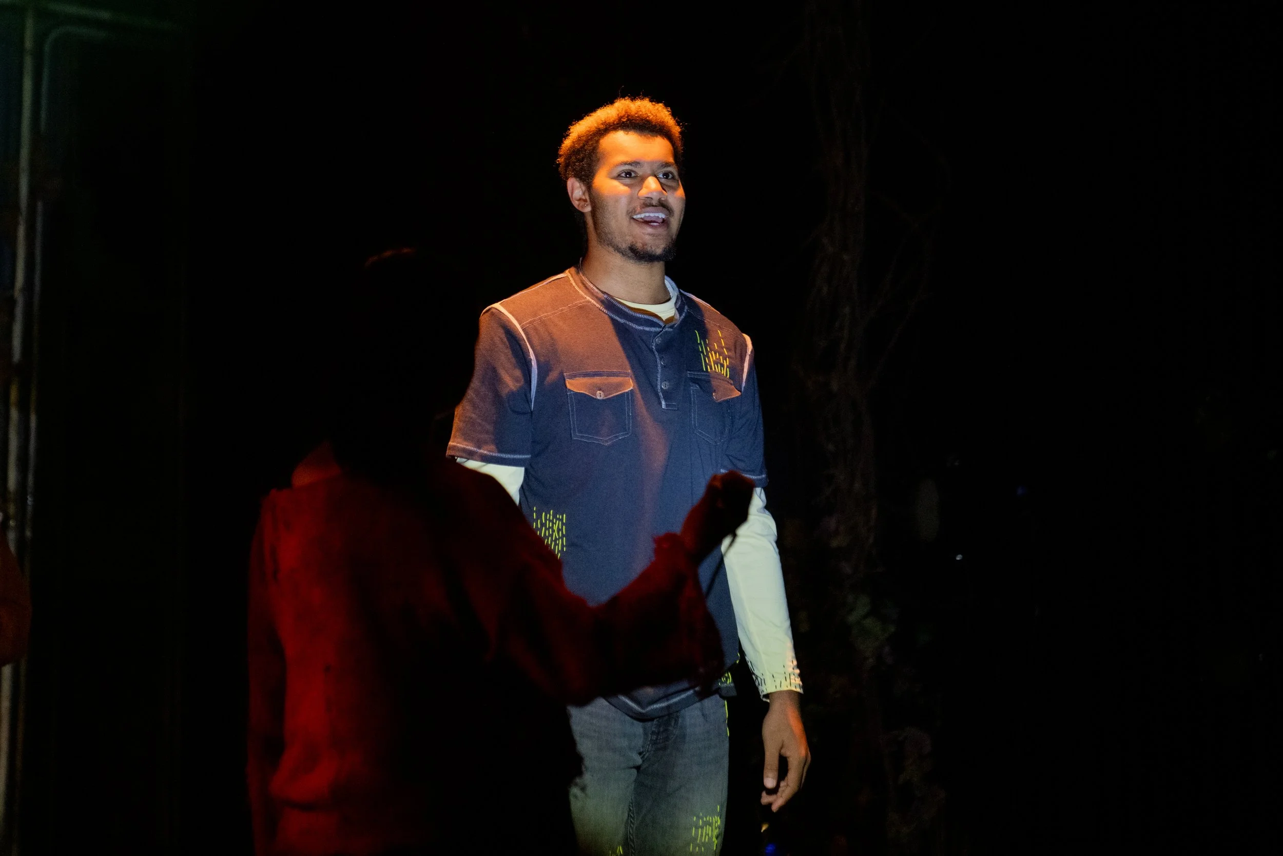 A young man with curly hair and a beard, wearing a dark short-sleeved shirt over a long sleeve shirt, standing on an outdoor stage at night, illuminated by stage lights.
