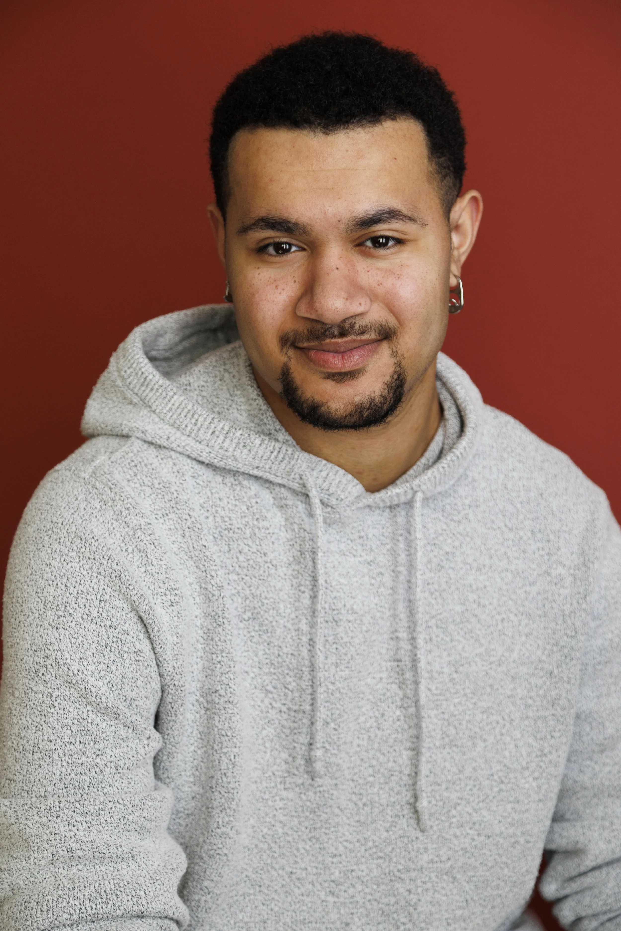 A young man with short dark curly hair, a goatee, and earrings, wearing a light gray hoodie, smiling in front of a solid reddish-brown background.