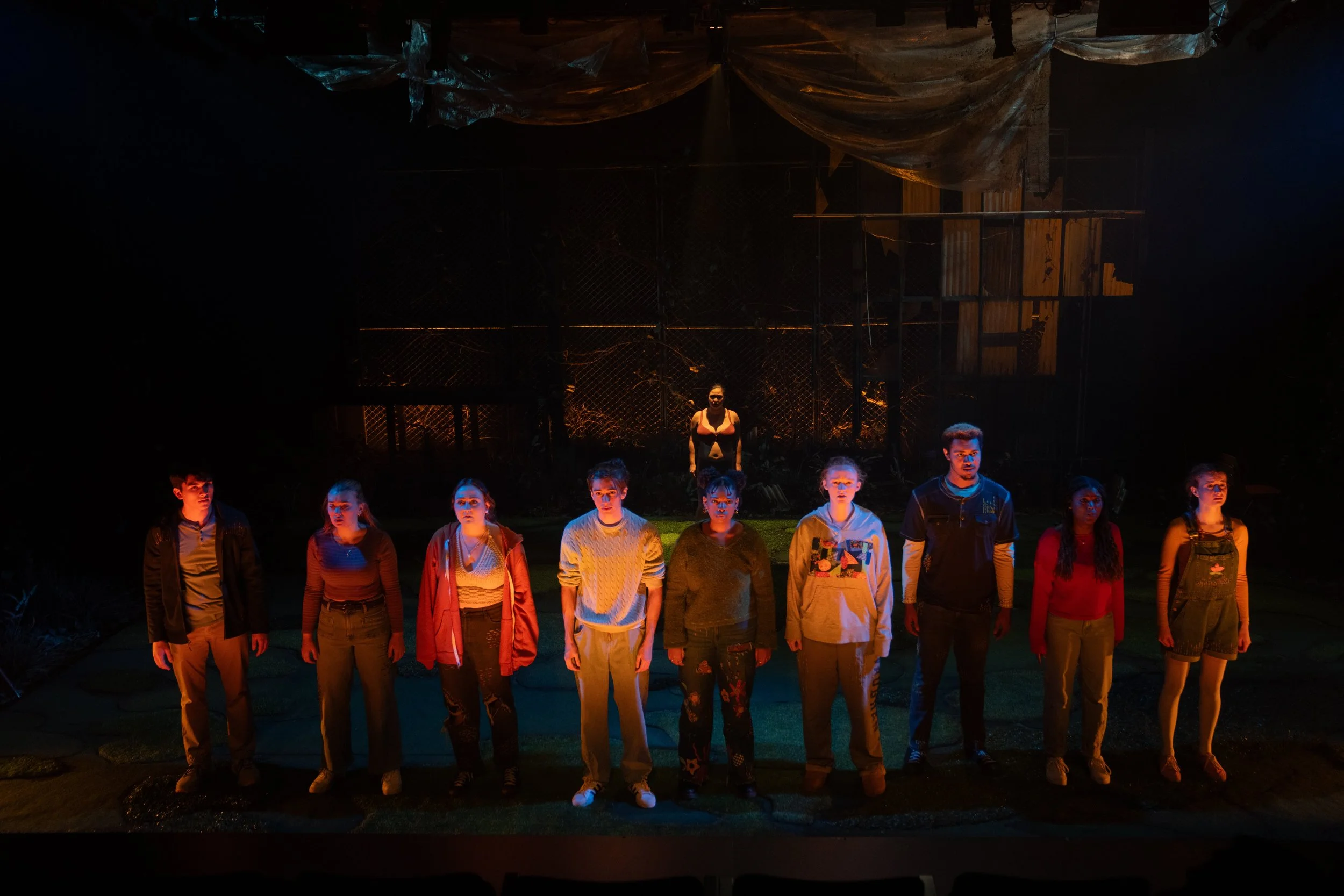 A group of eleven young actors on stage, standing in a line with a woman at the back center. The stage is dark with dramatic lighting highlighting the actors' faces, set against a backdrop resembling a chain-link fence and construction materials.