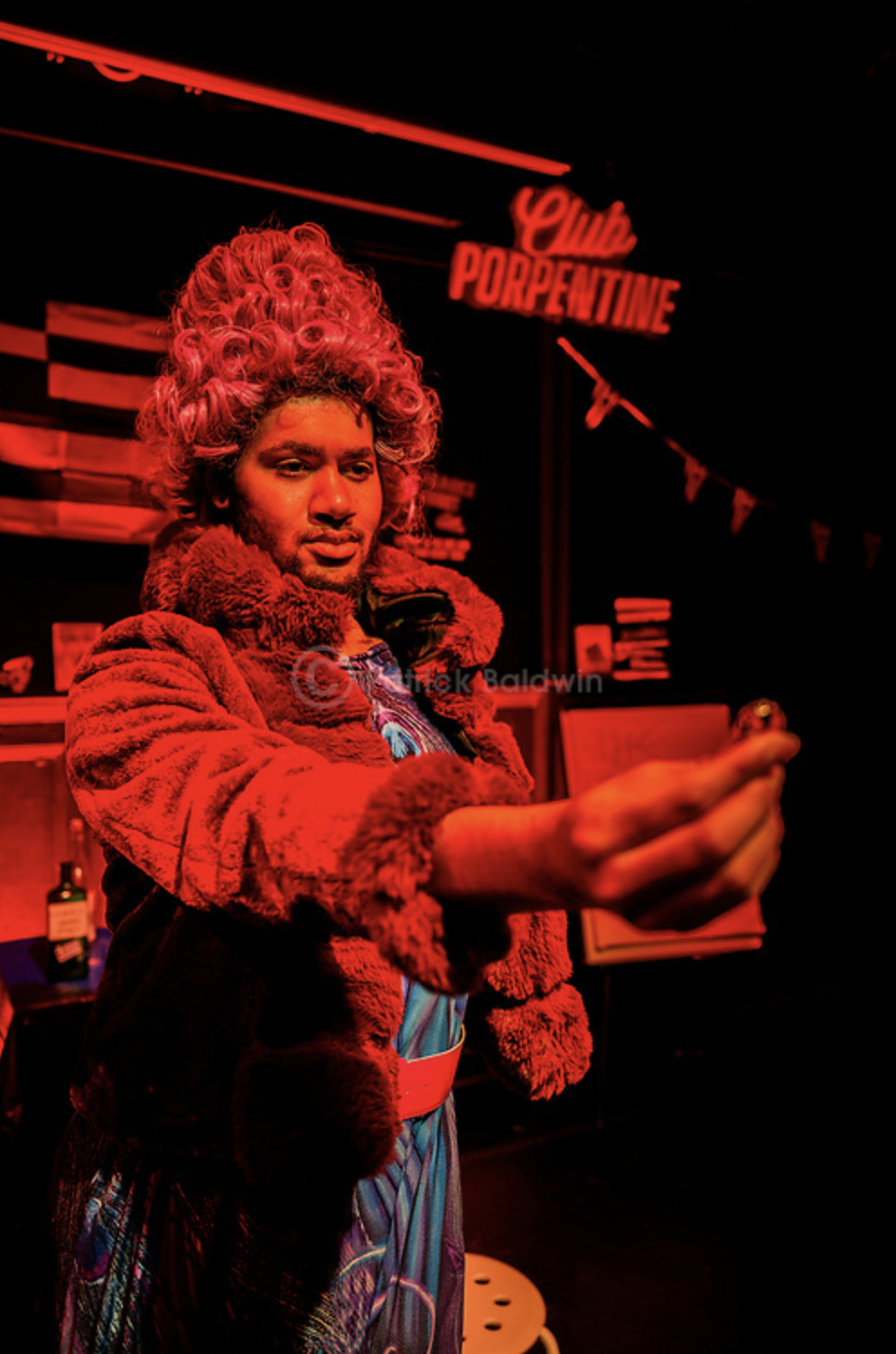 A man dressed in a flamboyant costume with a curly wig, fur coat, and colorful clothing, standing in a dimly lit club with neon signage that says 'Club Porpentine' in the background.