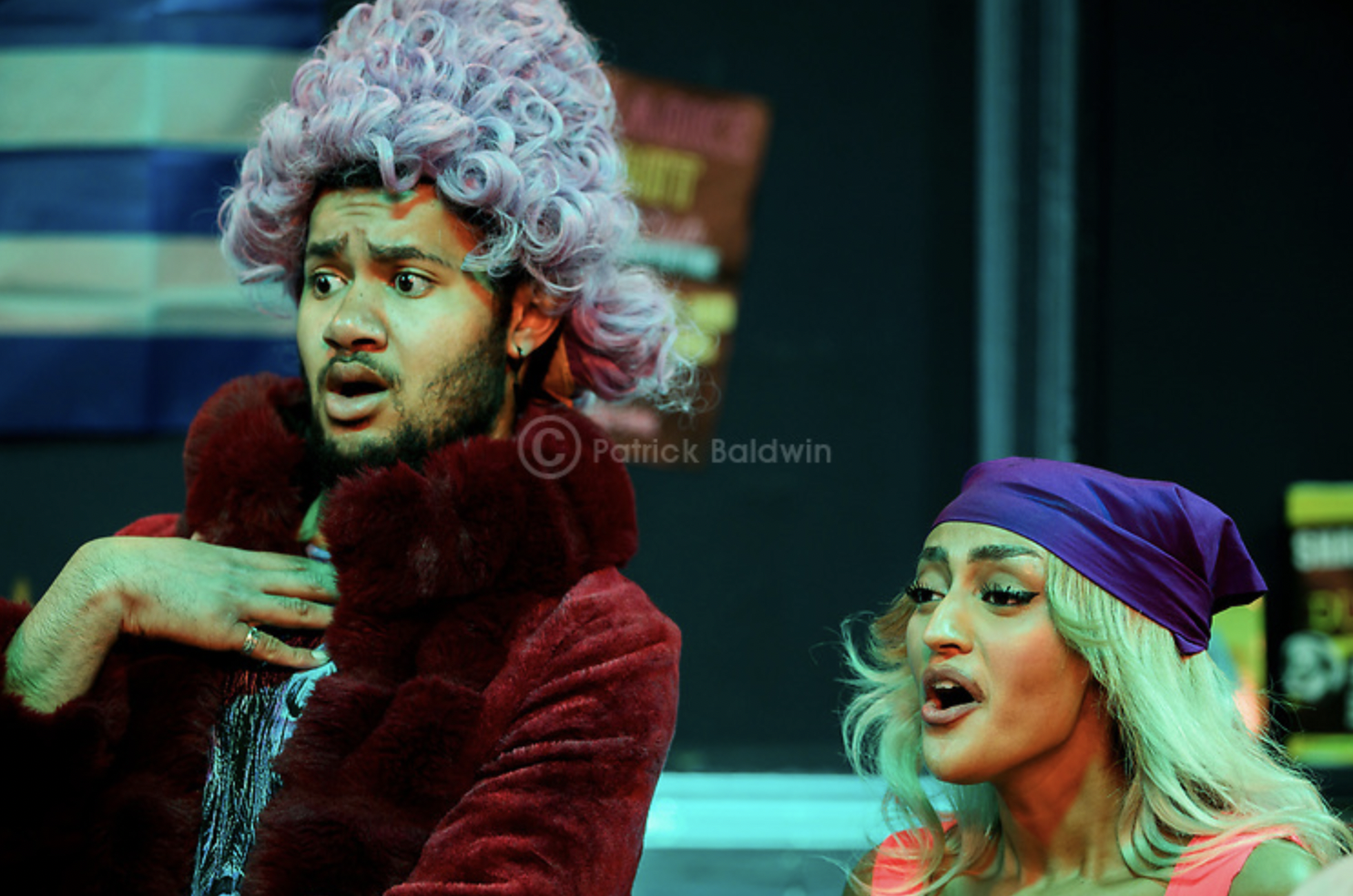 A man with a surprised expression wearing a curly wig, red fur coat, and holding his chest, and a woman with a bandana on her head, blonde hair, and a surprised expression, standing in a colorful indoor setting.