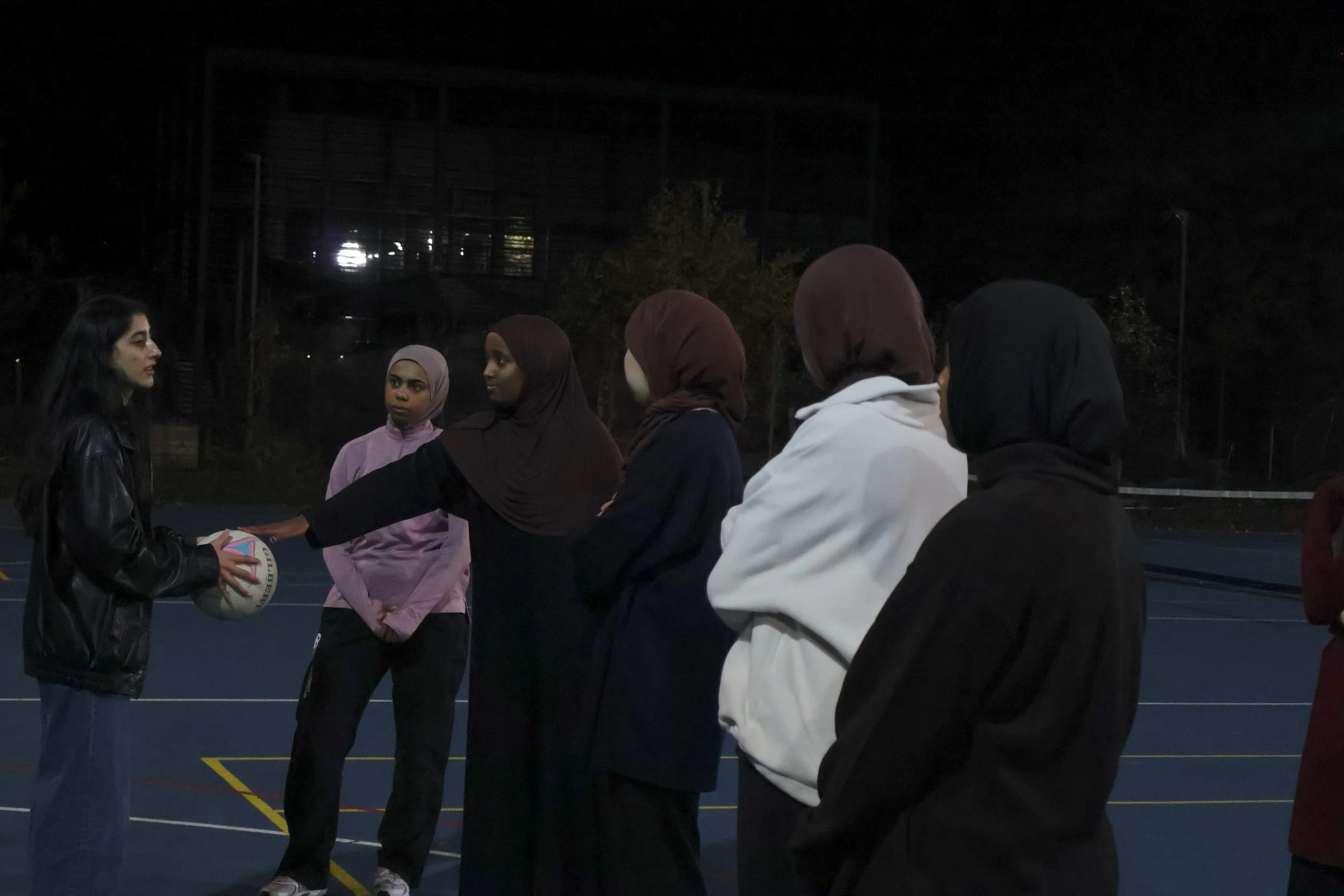 Night scene of women playing basketball on an outdoor court, with some women standing and reaching up, and others watching, surrounded by a tall fence, with a brick building and people in the background.