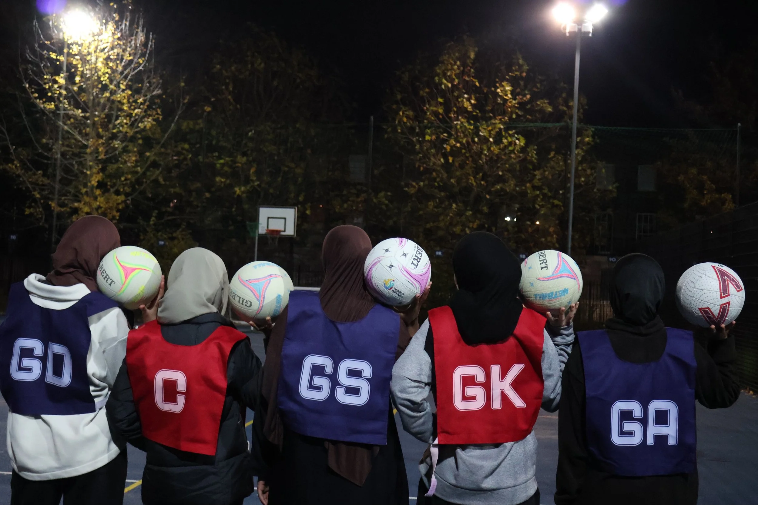 A woman with long dark hair coaching a group of women in hijabs on a sports court at night, holding a rugby ball.