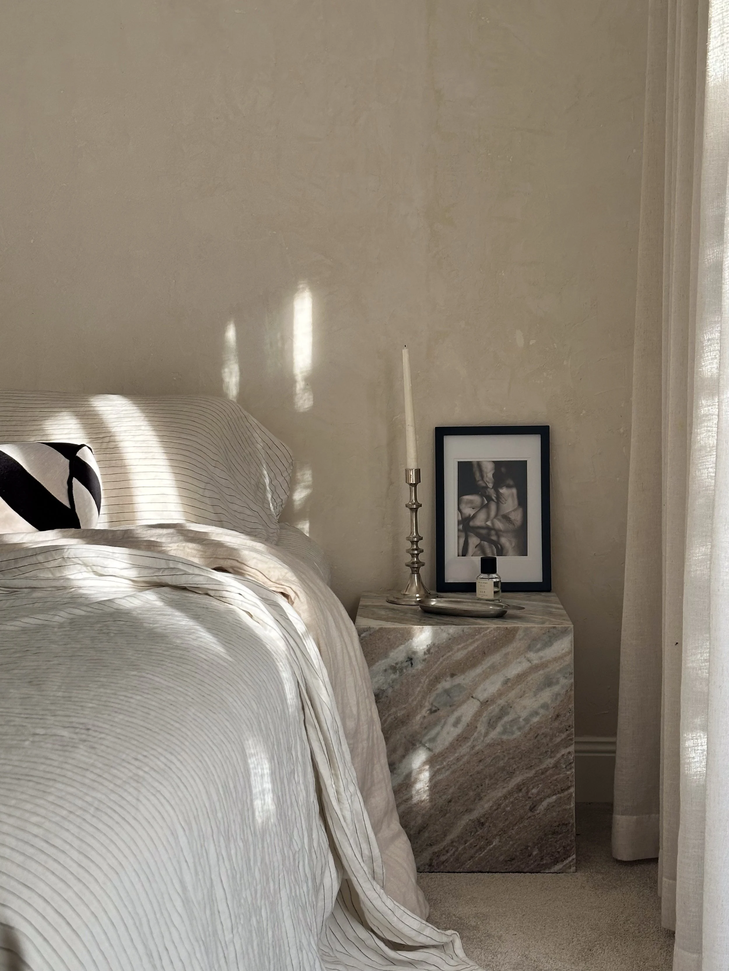 A bedroom corner with a bed, a marble nightstand, a framed black-and-white photo, a candlestick, and a small bottle of perfume. Sunlight creates shadows on the wall.