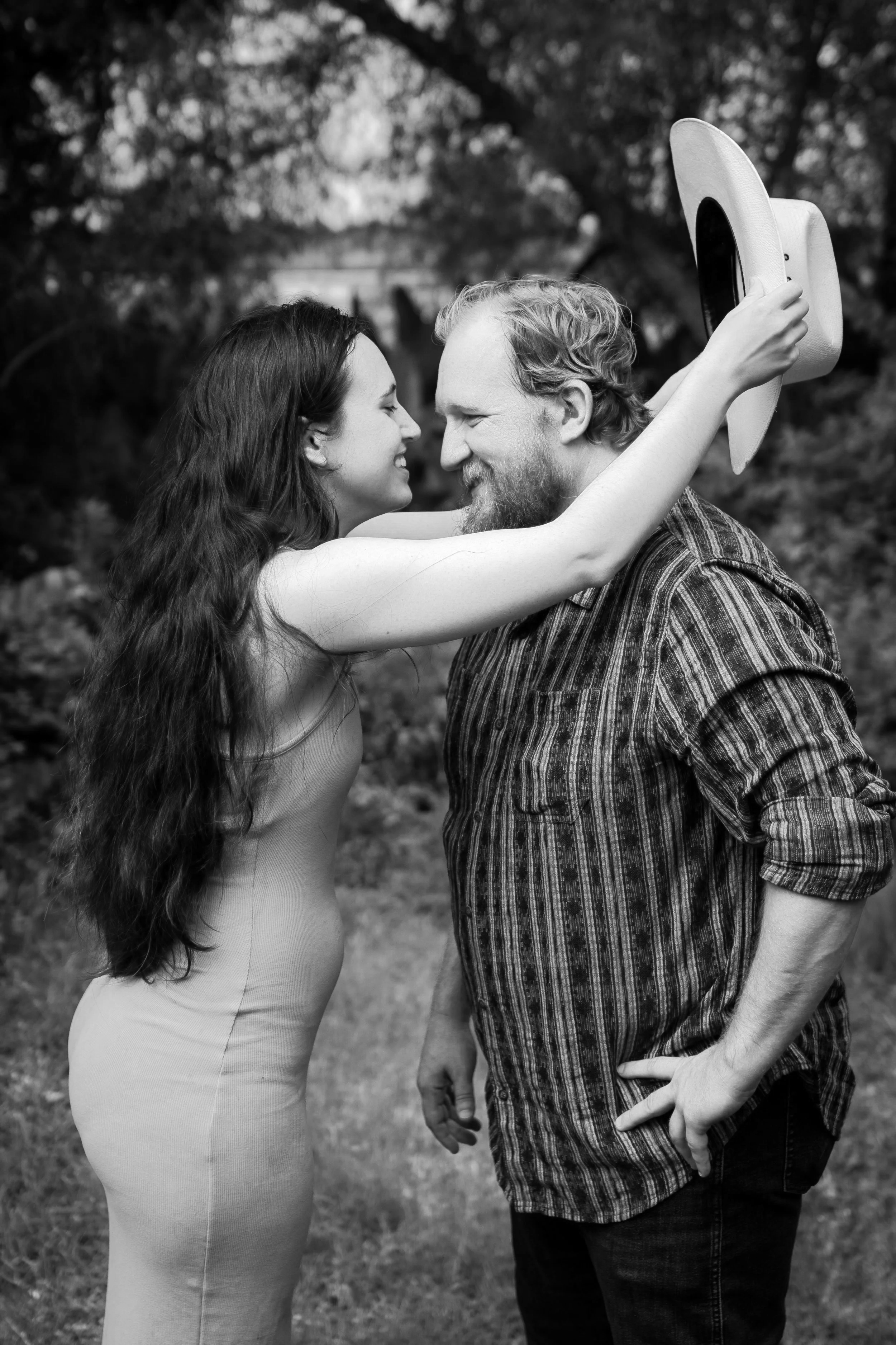 A black and white photo of a woman and a man standing close together outdoors, smiling and about to kiss. The woman is holding a boomerang in the air. Trees are visible in the background.