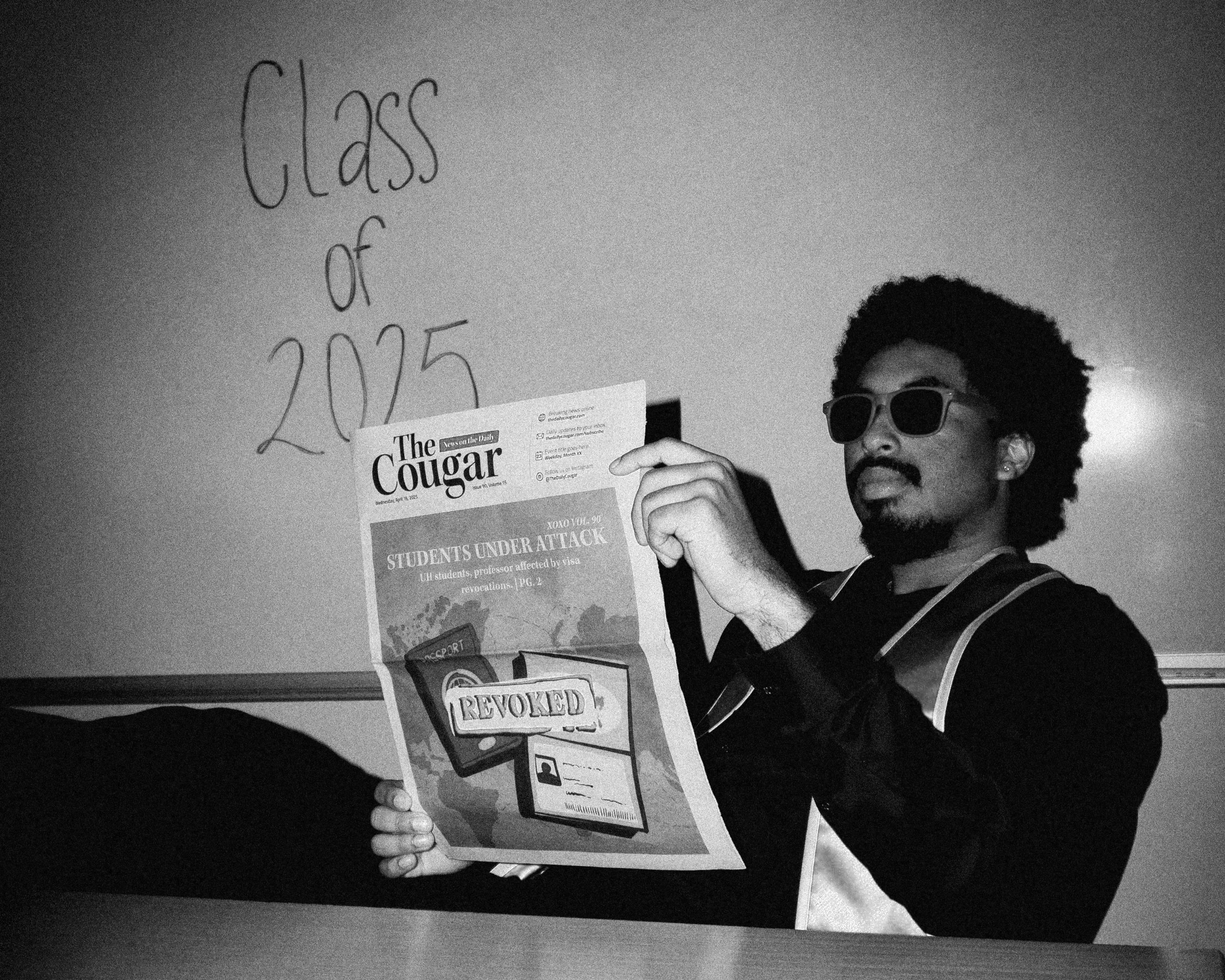 A person with curly hair and sunglasses holding a newspaper titled 'The Cougar,' which has headlines about students under attack and revoking rights, standing in front of a wall with handwriting that says 'Class of 2025.'