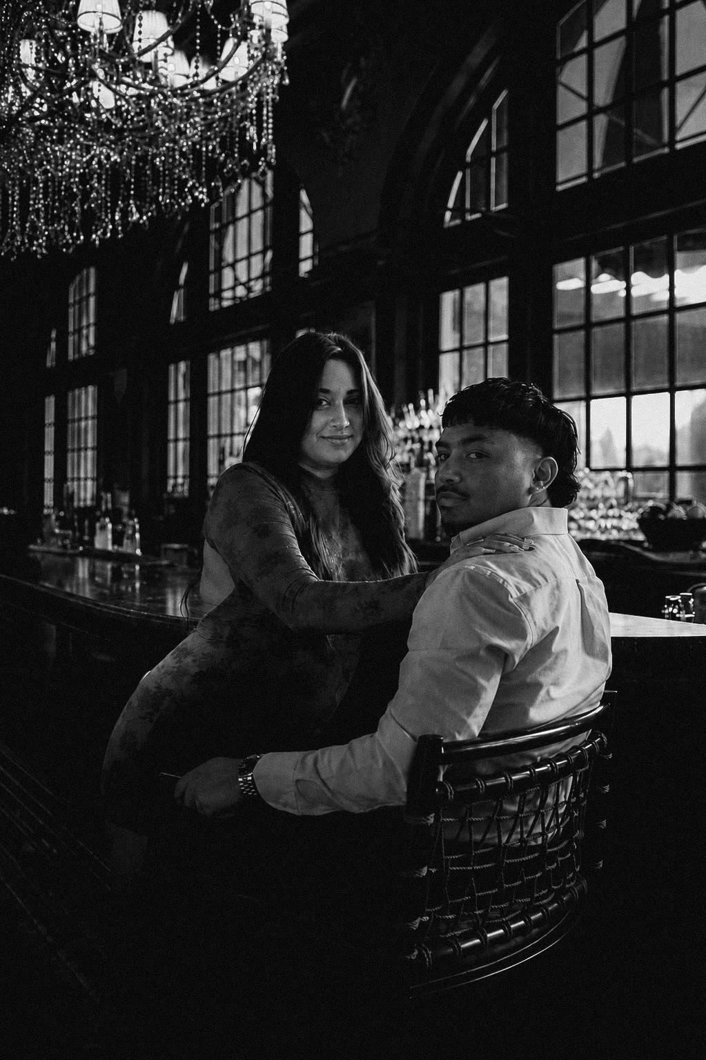 A black-and-white photograph of a woman and a man sitting inside a dimly lit, stylish bar or restaurant. The woman is leaning on the bar counter with a subtle smile, and the man is sitting on a chair, looking towards the camera. Large windows in the background allow natural light to filter in, and a chandelier with hanging beads decorates the ceiling.