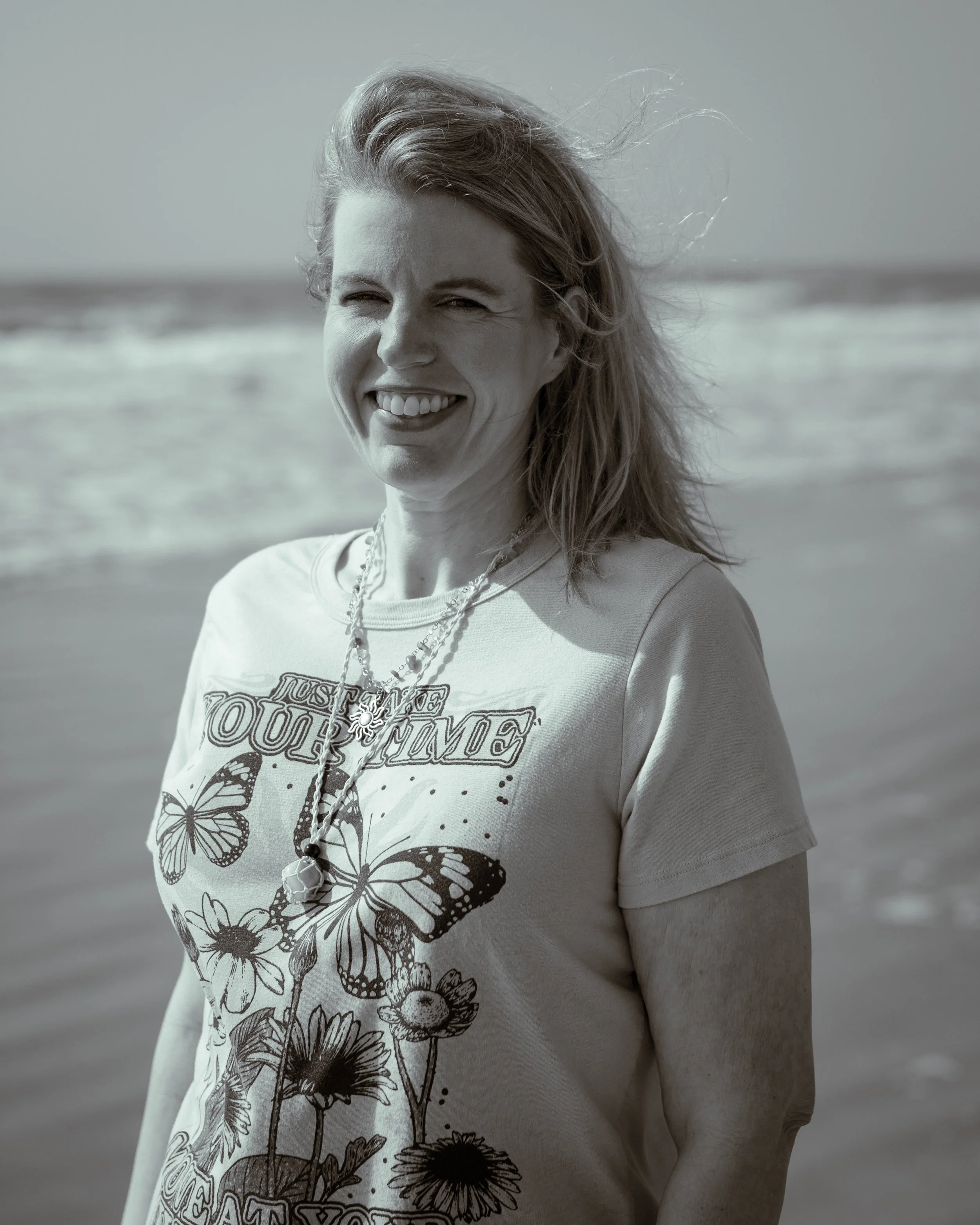 A woman smiling at the camera at the beach with wind-blown hair, wearing a T-shirt with floral and butterfly prints.
