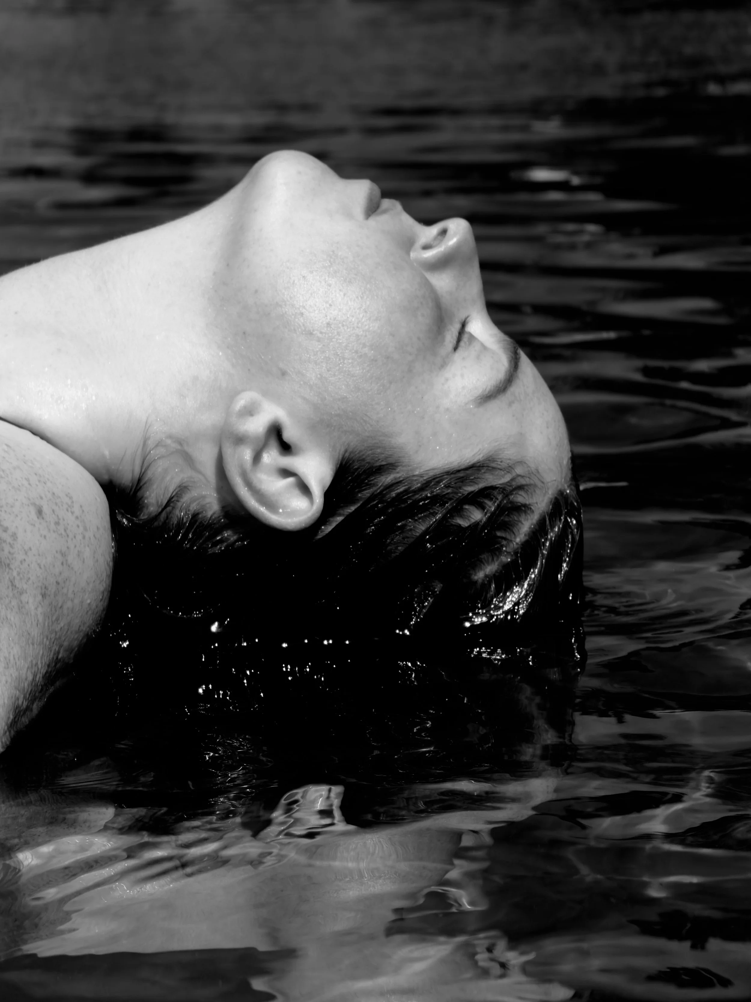 A black and white close-up photo of a person floating face-up on water, with their face and nose above the water and their wet hair submerged.