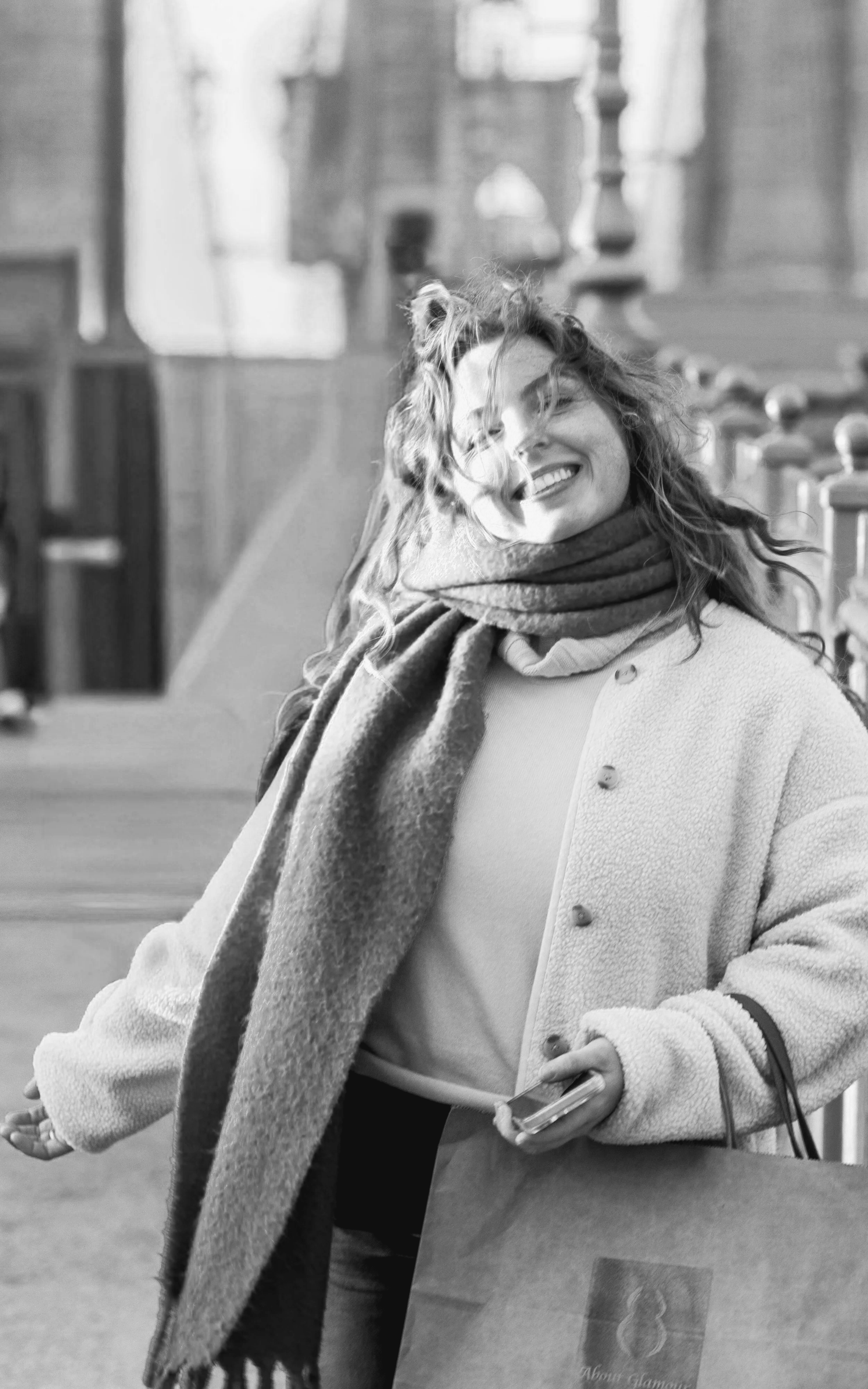 A woman smiling with closed eyes, wearing a coat and scarf, holding a purse and phone, walking outdoors with blurred buildings in the background.