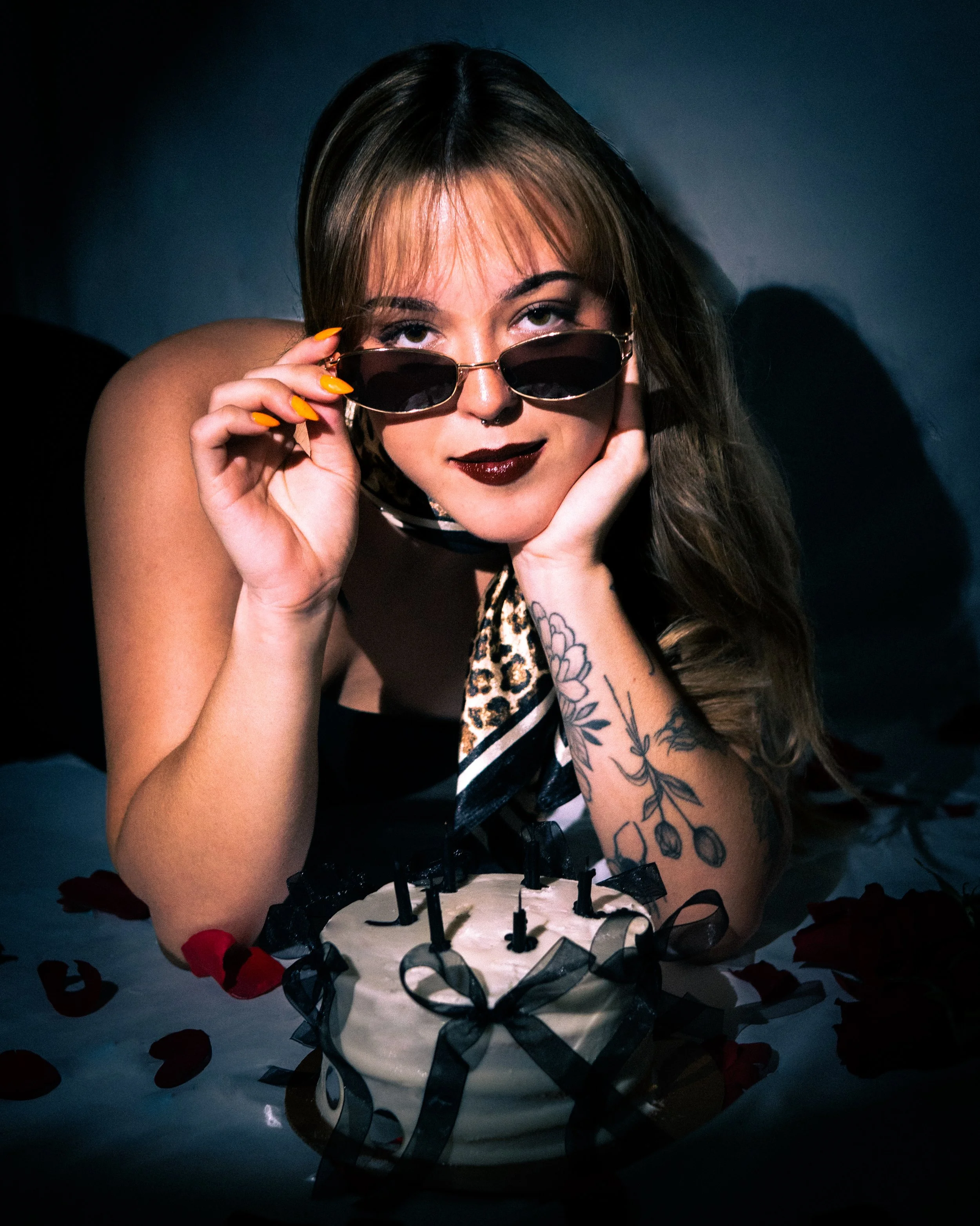 A woman with dark lipstick, sunglasses partly lowered, intense gaze, tattoos on her arm, and styled hair, holding a finger near her face surrounded by rose petals. She is sitting at a table with a decorated cake.