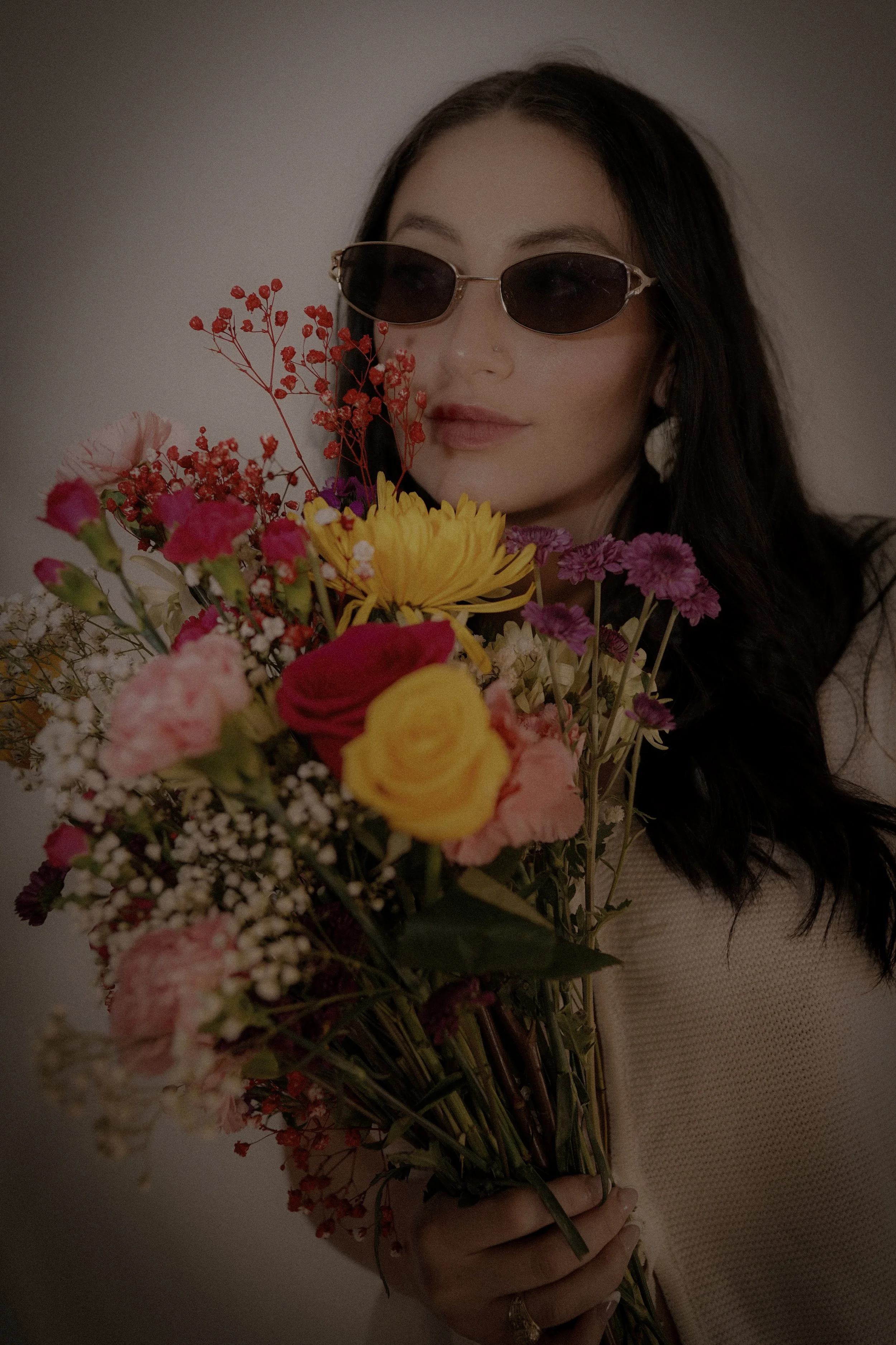 A woman with dark hair wearing sunglasses and a beige top is holding a large colorful flower bouquet
