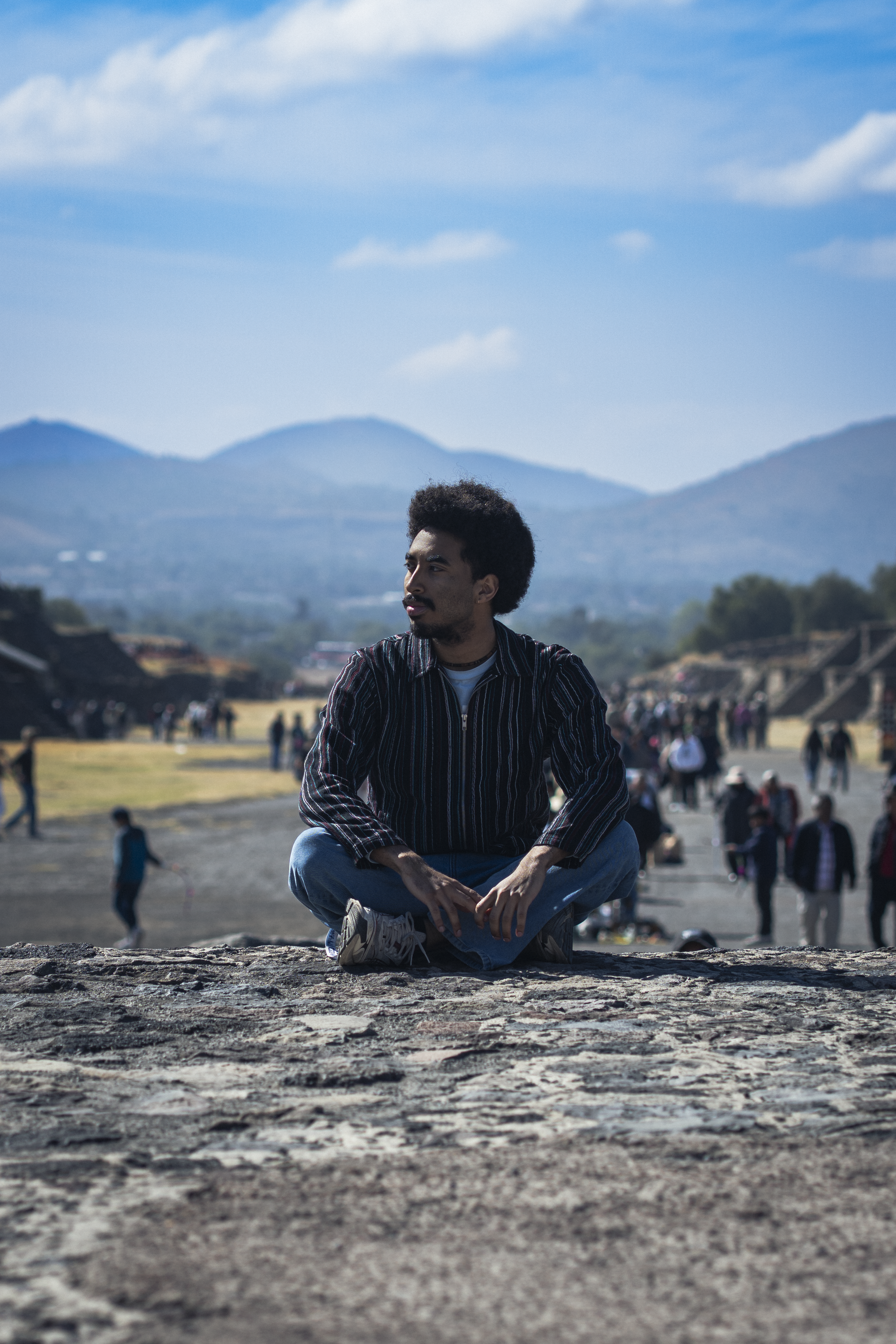 A young man with curly hair and a beard is sitting cross-legged outdoors on a rock, with a crowd of people and traditional houses in the background. Mountains and a partly cloudy sky are visible in the distance.