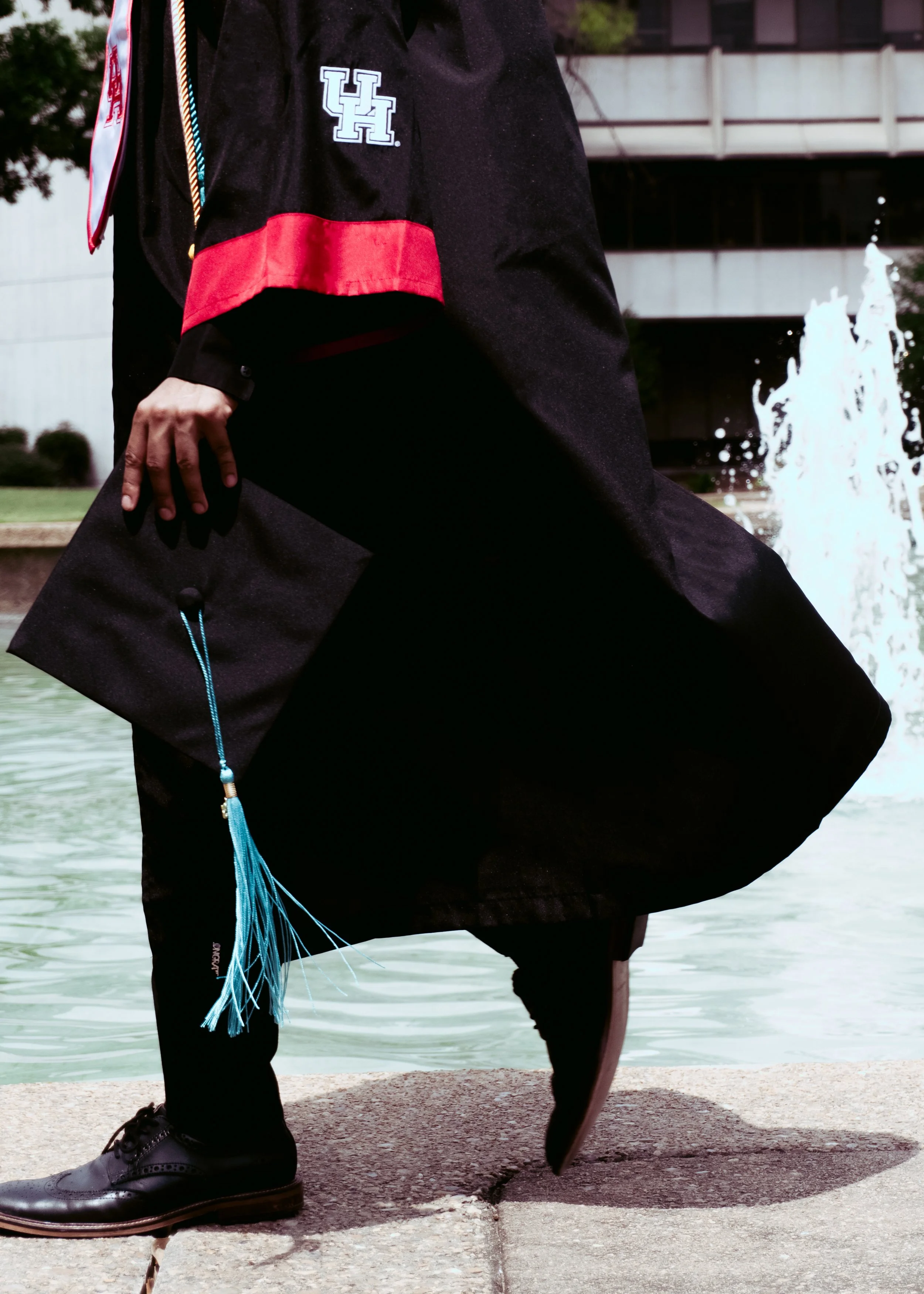 Close-up of a person wearing a graduation gown and cap, holding a diploma, standing near a fountain with water sprays, background includes modern buildings.