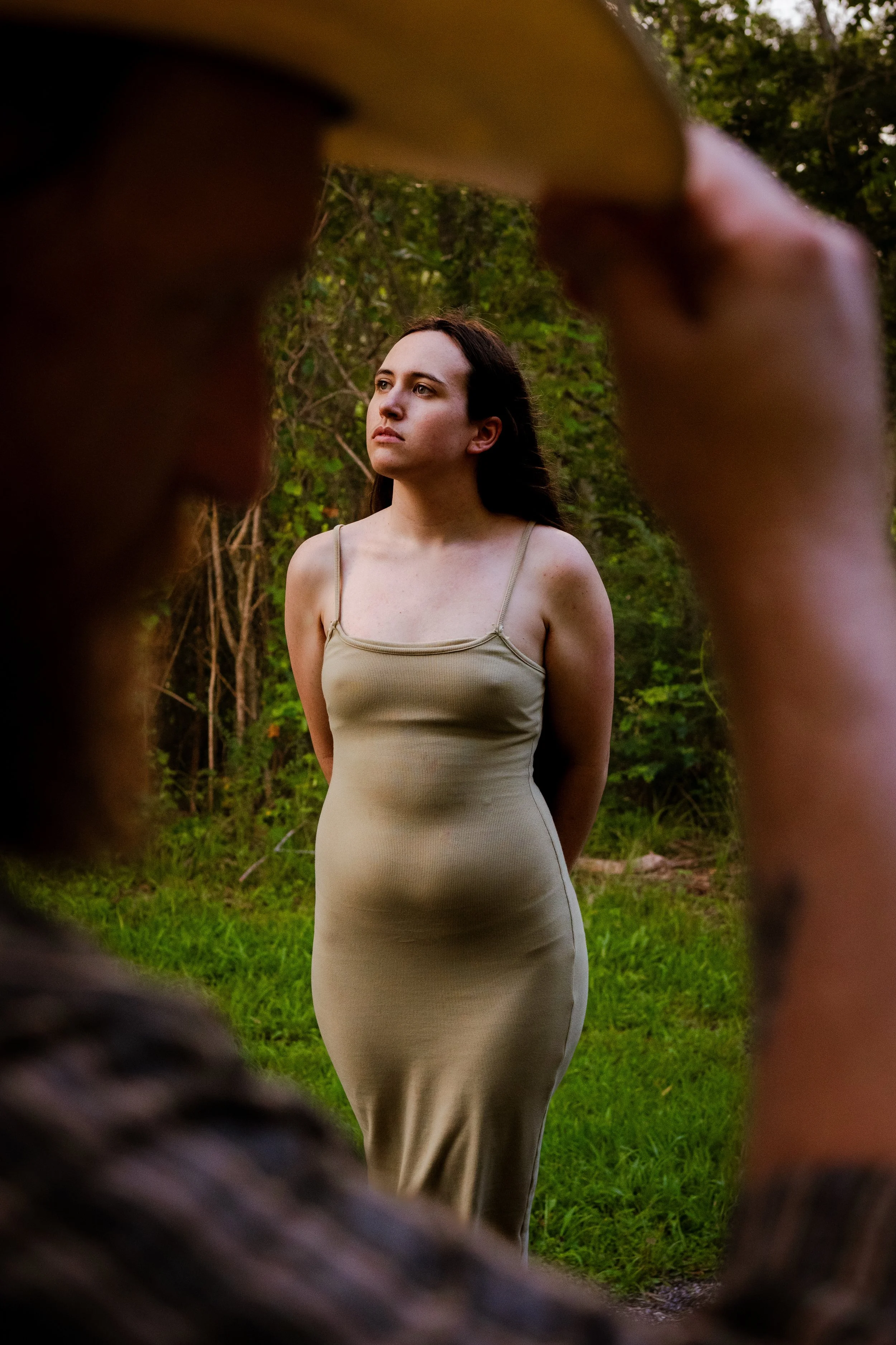 A woman with dark hair wearing a beige sleeveless dress stands outdoors in a wooded area, framed by the blurred outline of a person's foot in the foreground.
