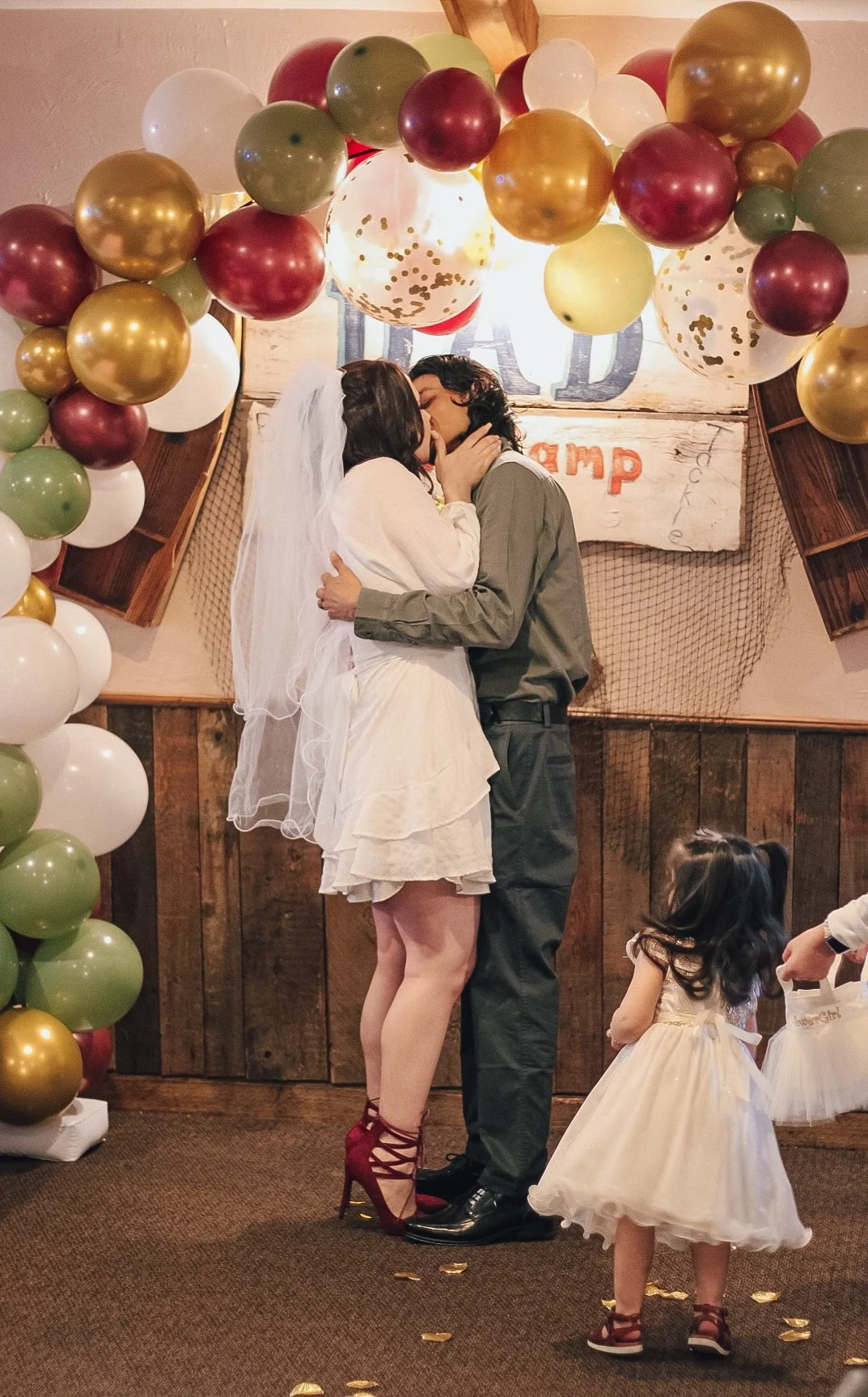 A couple kissing during a wedding ceremony, with a woman in a white dress and red heels, and a man in black pants and a grey shirt. Two young girls in white dresses with gold bows are nearby, one holding a small bag. The scene is decorated with multi