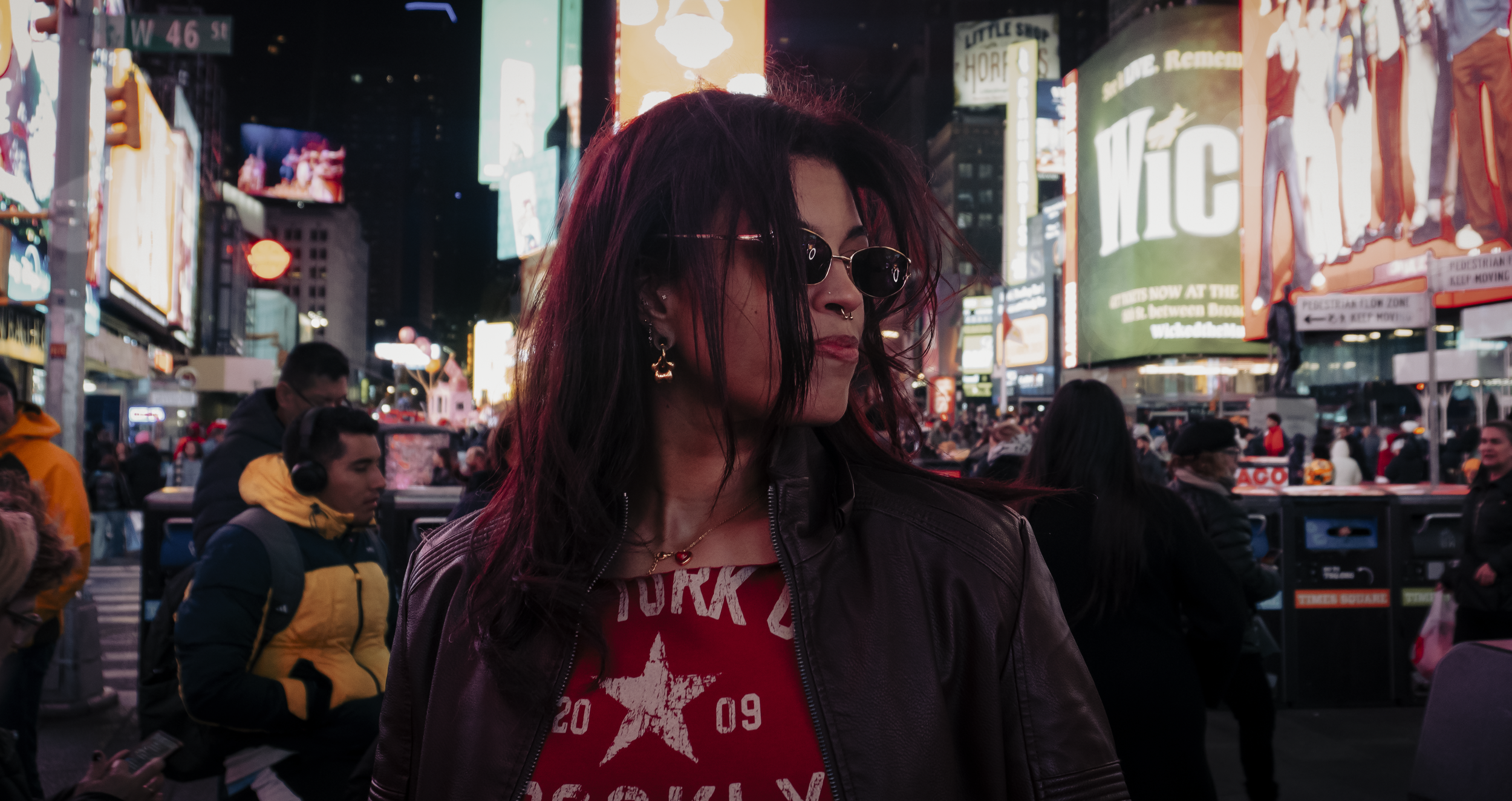 A woman with dark red hair, wearing sunglasses, earrings, and a black leather jacket, standing in a crowded city square at night with bright billboards and electronic advertising signs in the background.