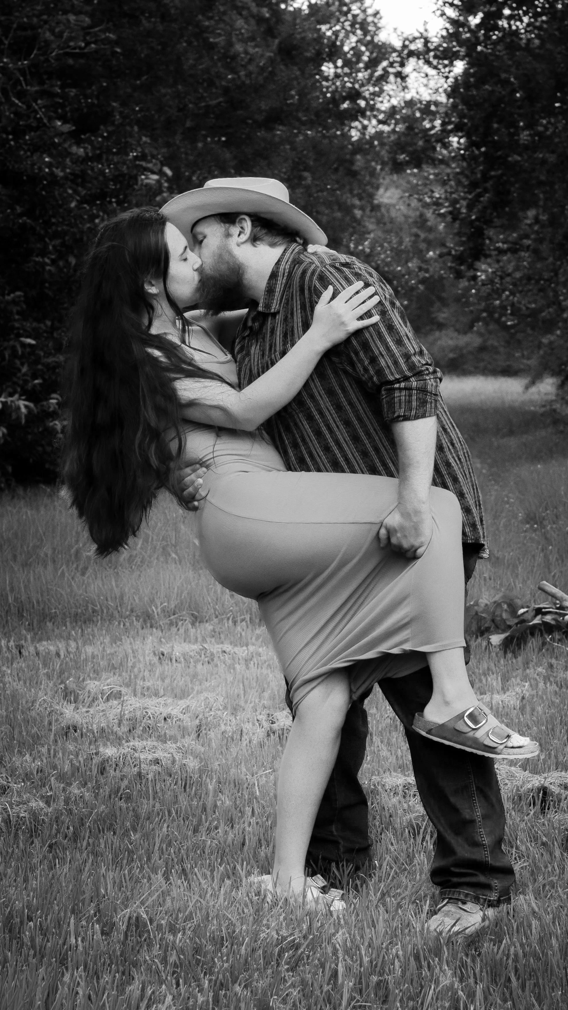 A man with a cowboy hat carrying a woman in a dress, sharing a kiss outdoors in a grassy area with trees in the background.