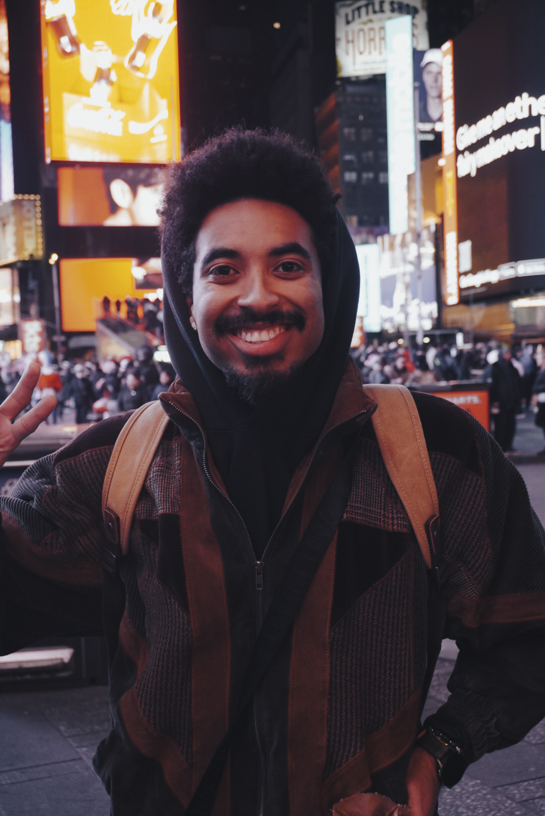 A smiling man with a backpack at night in Times Square, New York City, illuminated by bright billboards and city lights.