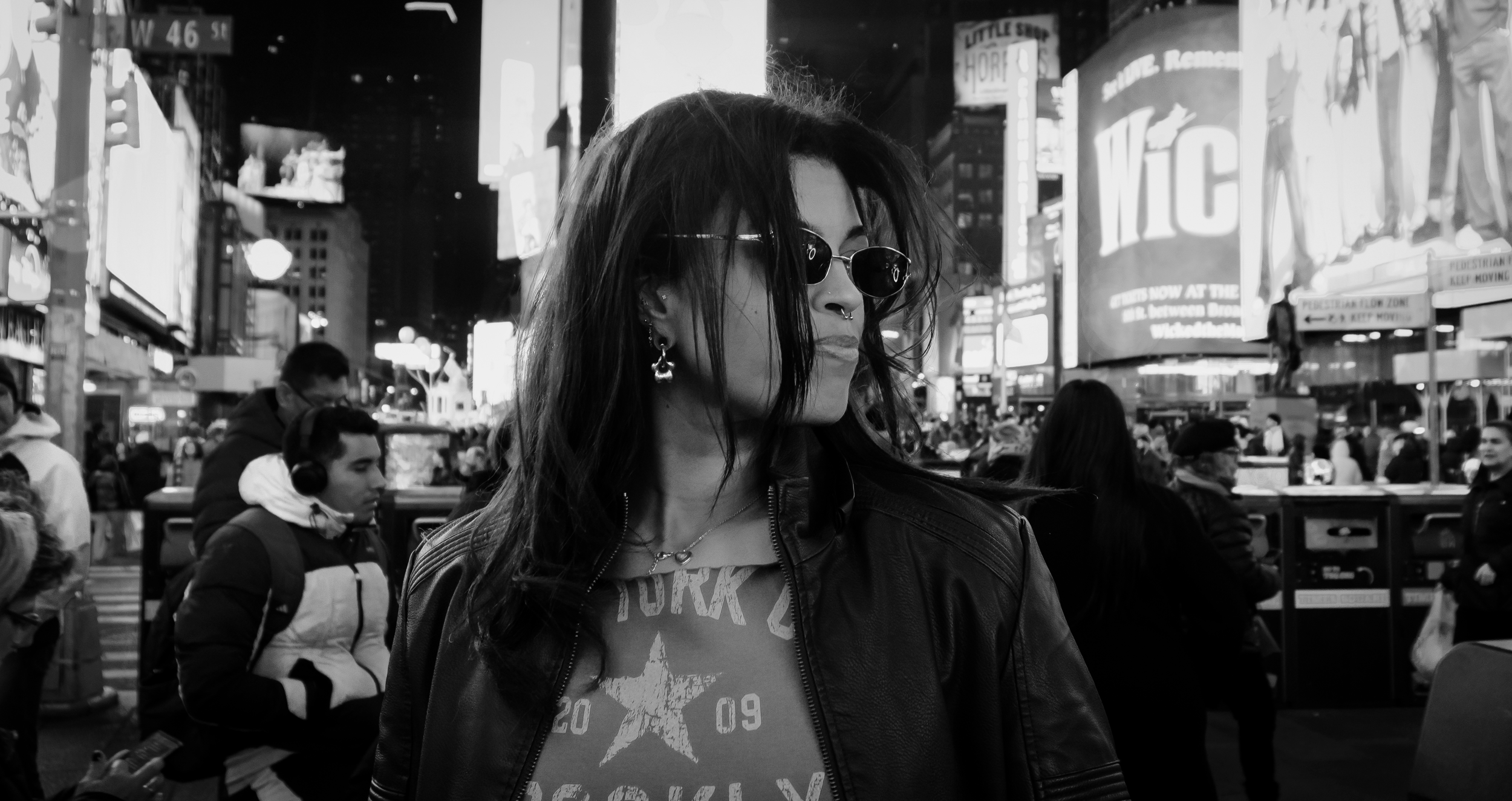 A woman with dark hair, wearing sunglasses, earrings, and a leather jacket, standing in Times Square, NYC at night surrounded by a crowd and illuminated billboards.
