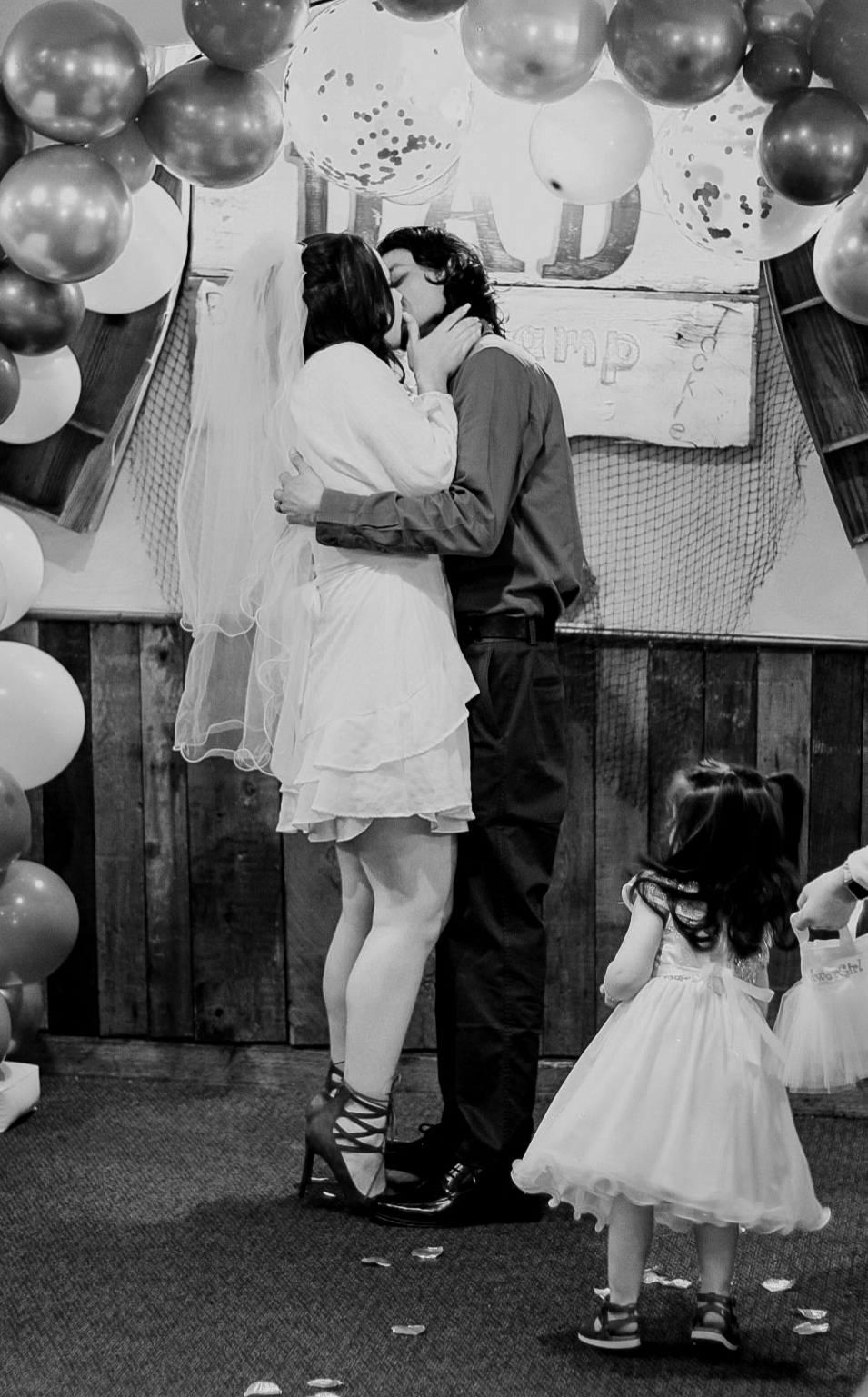 A couple kissing during a wedding reception, with a young girl in a dress watching nearby, surrounded by balloons and wedding decorations.
