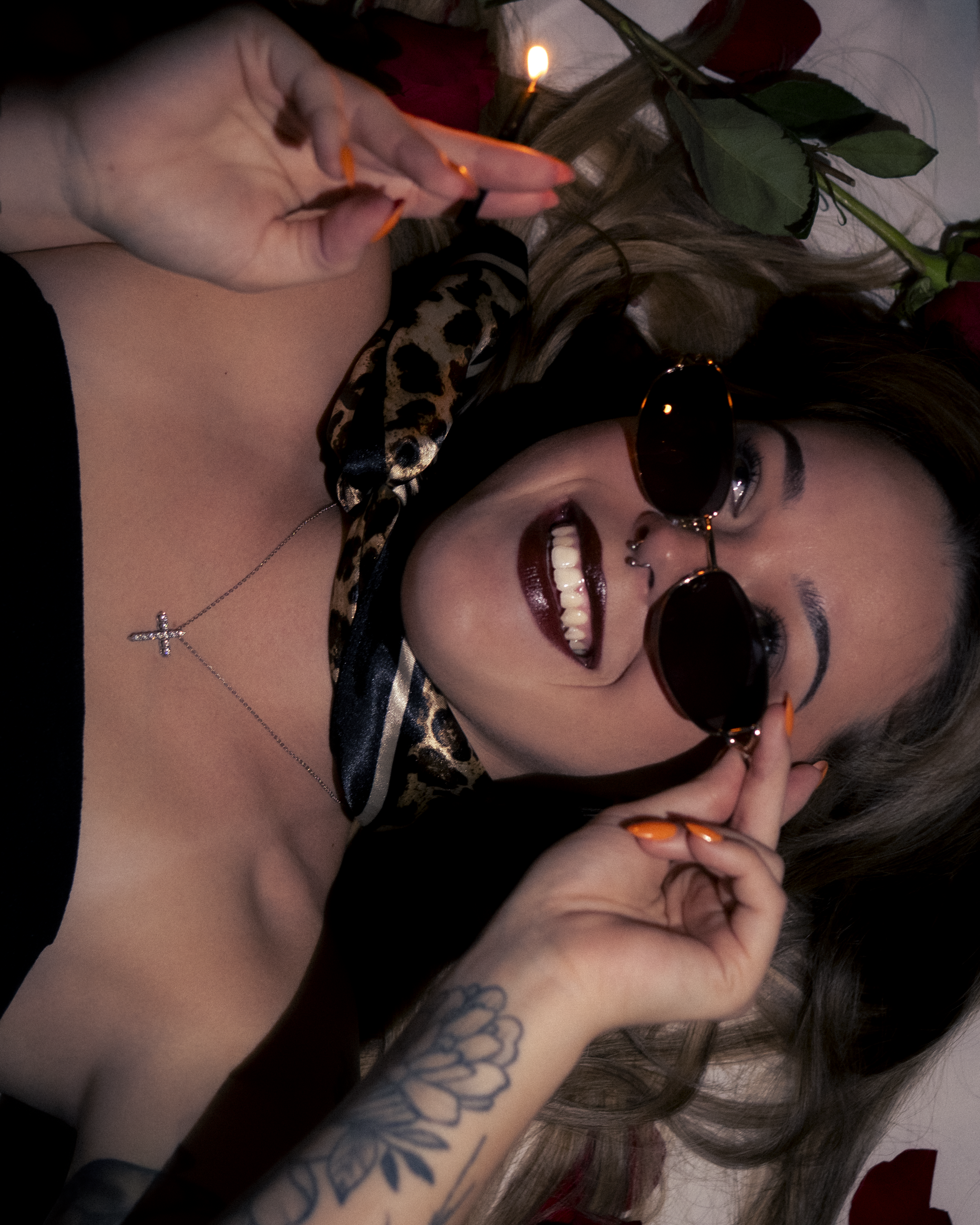 A woman with long hair, wearing sunglasses and a black top, smiling and showing her teeth. She has tattoos on her arm and a cross necklace. She is holding a lit matchstick. There are roses on her hair, and the background is plain.
