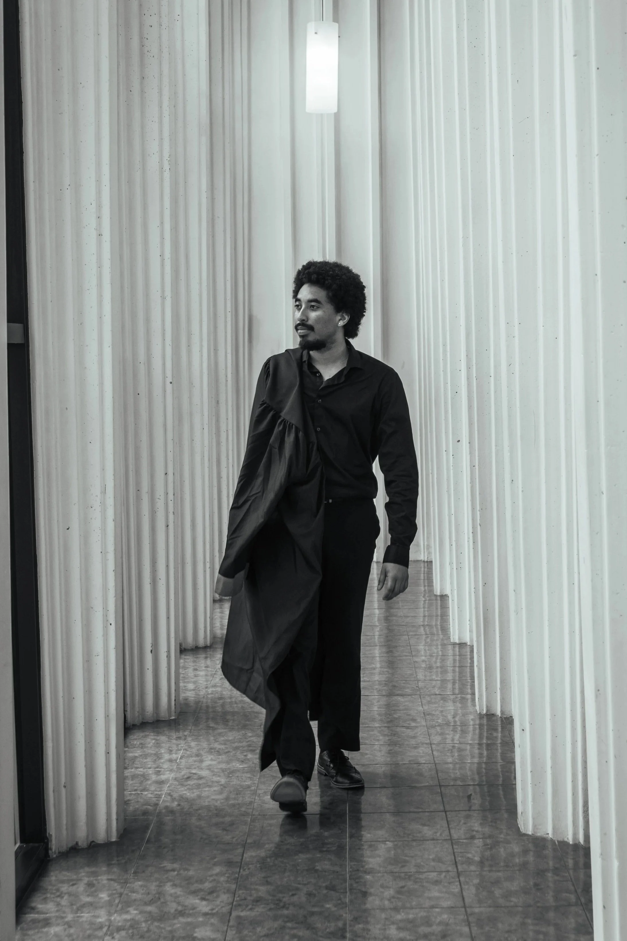A man in black clothing walking in a corridor with white, vertically-striped curtains on either side, tiled floor, and a hanging light. He's holding a black jacket over his shoulder.