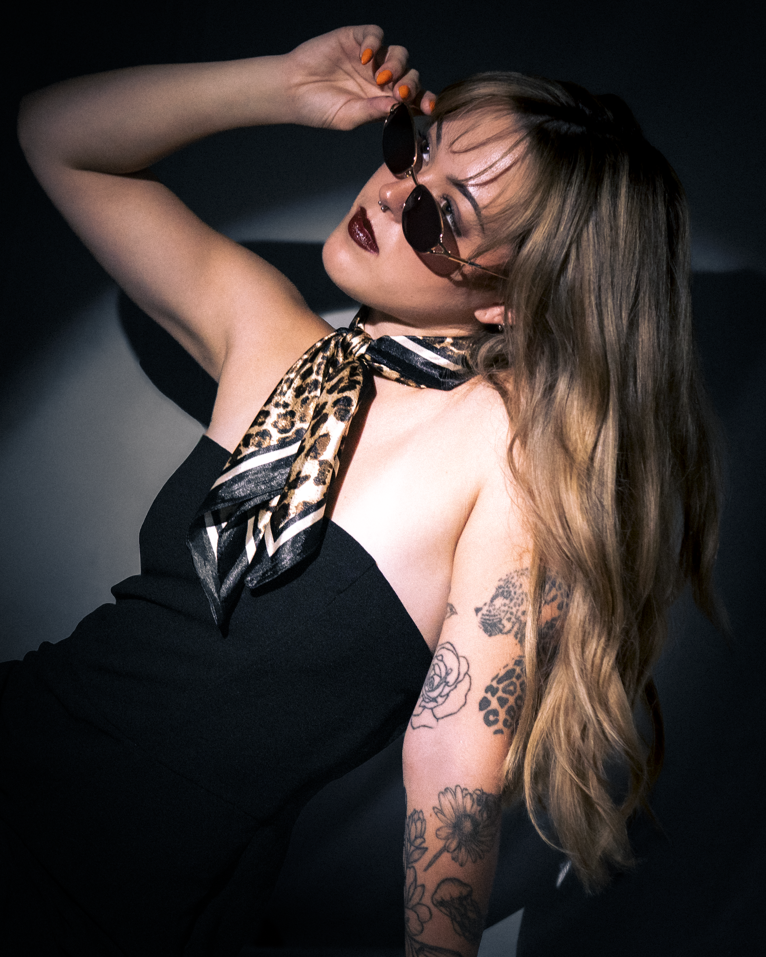 A woman with long, wavy hair and tattoos on her arm is holding sunglasses up to her face, looking off to the side with a confident expression.