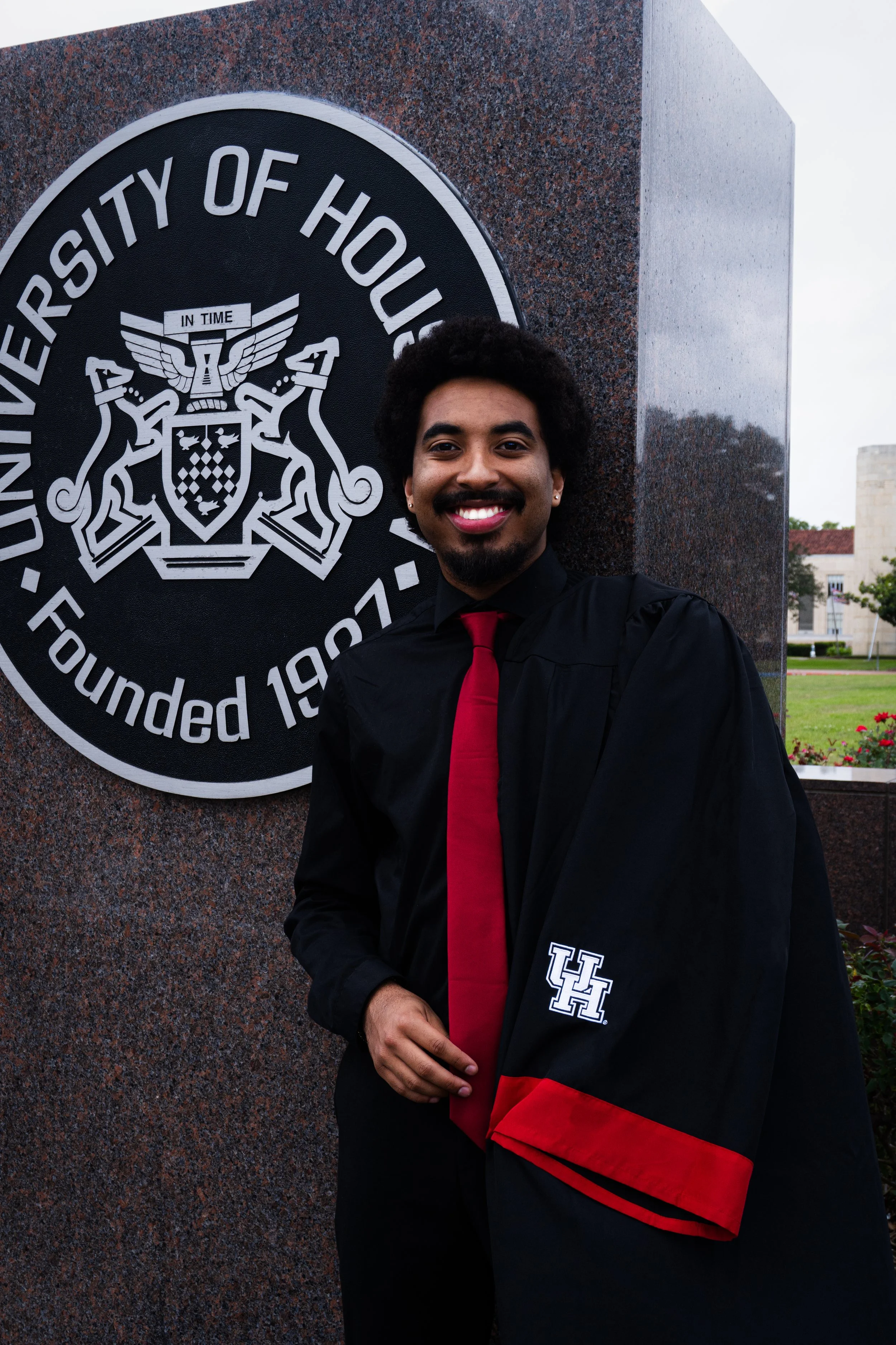 Young man in graduation gown and cap standing next to a university emblem, smiling outdoors on a cloudy day.