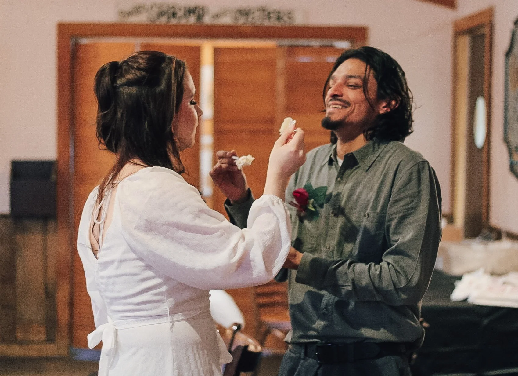 A man and woman sharing a romantic moment indoors, with the woman feeding the man cake and the man smiling, on a warm occasion.