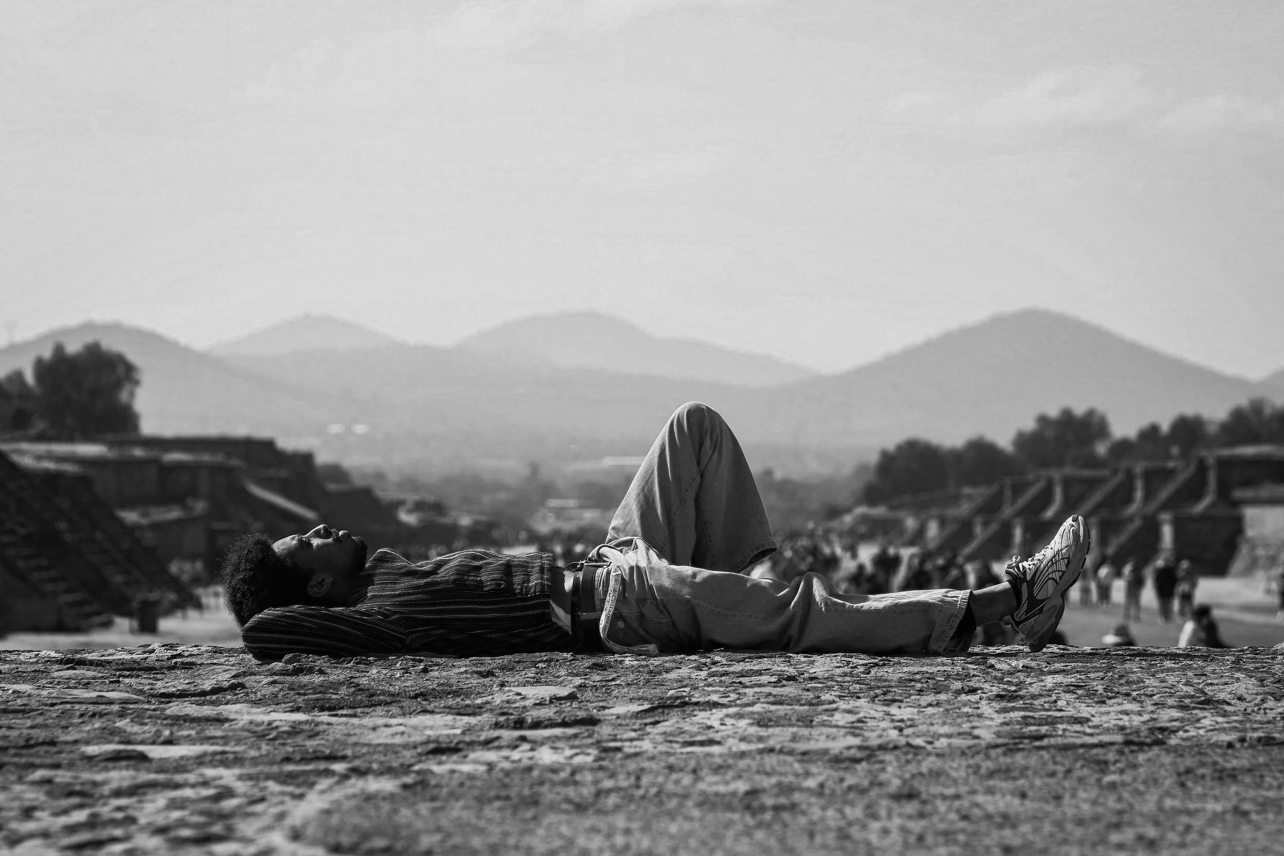 A person lying on their back with arms and legs relaxed on a ground surface with mountains and trees in the background, in black and white.